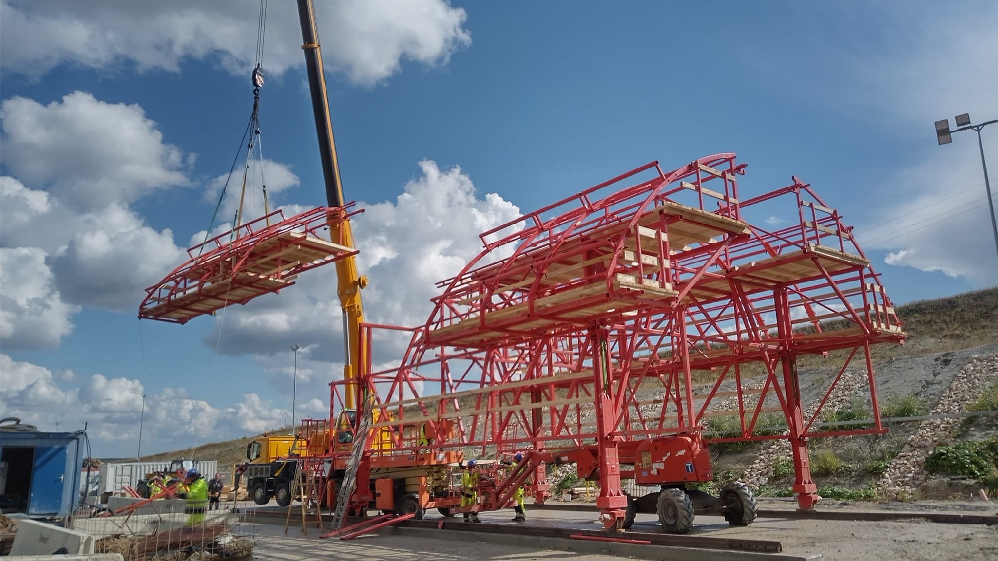 Construction workers assembling a large red steel structure using a crane on a construction site with cloudy sky.