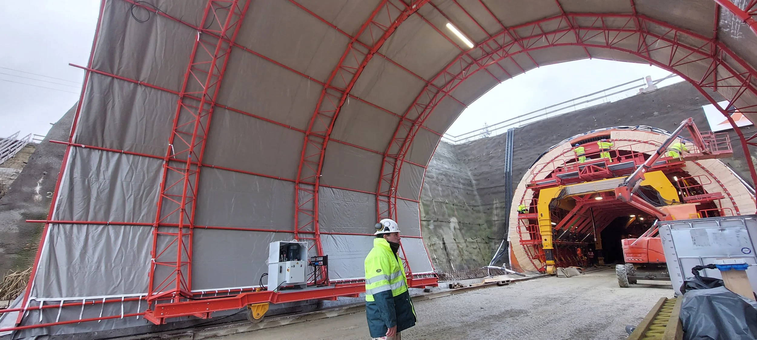 Construction workers in high-visibility jackets and helmets are working inside a tunnel being excavated, with the tunnel's entrance visible on the right and construction equipment around.