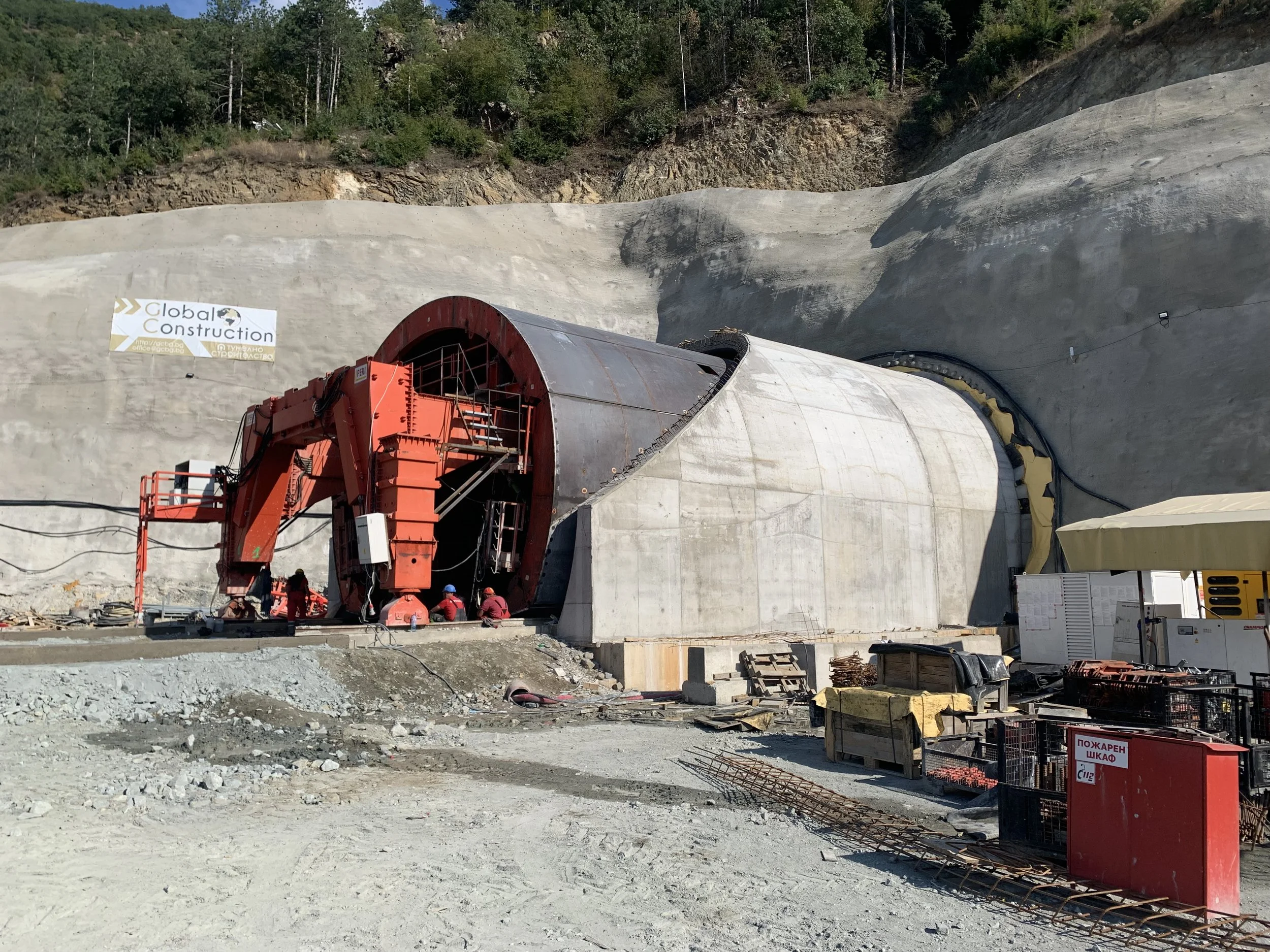 Construction site with a tunnel being built into a hillside. The entrance has a large red support structure and workers in red uniforms nearby.