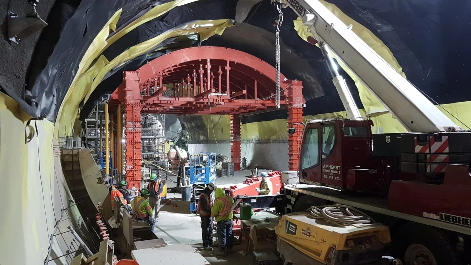 Construction workers in an underground tunnel with a large red arch structure, scaffolding, machinery, and construction equipment.