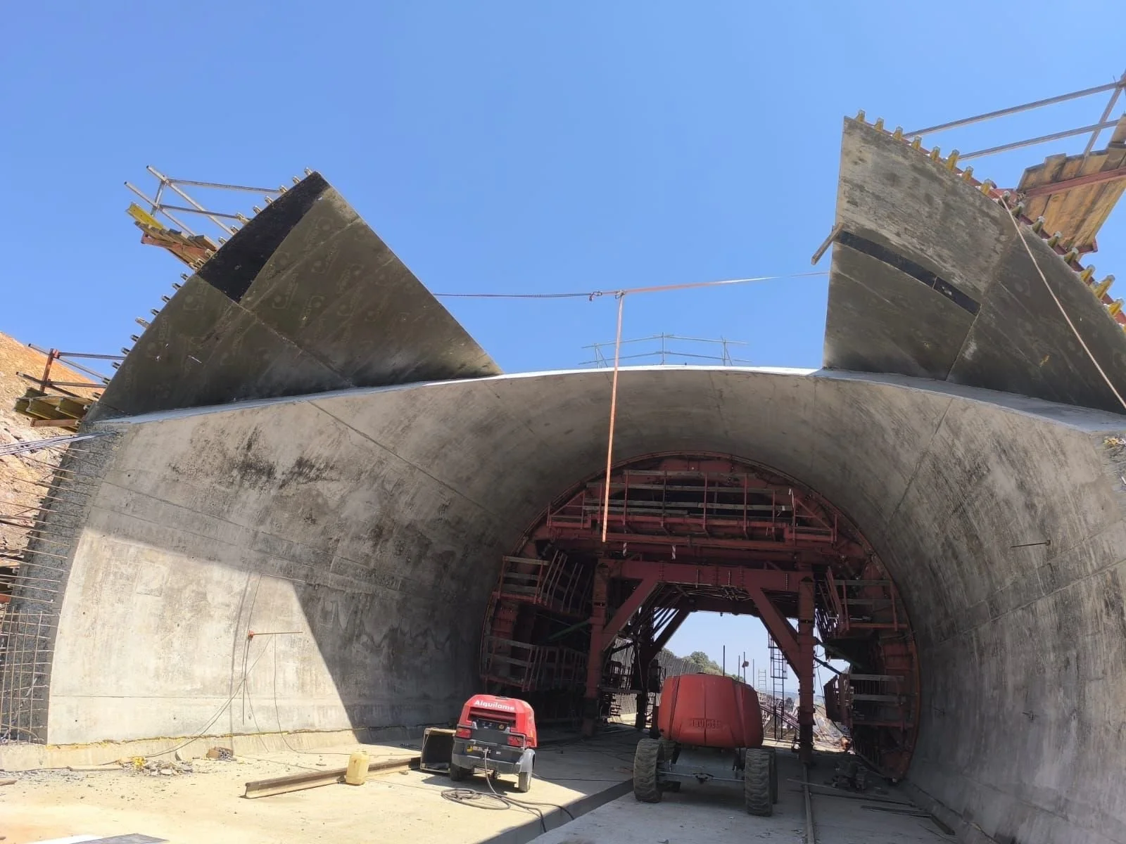 Under construction tunnel with equipment and scaffolding, partially completed concrete and steel framework, clear blue sky above.