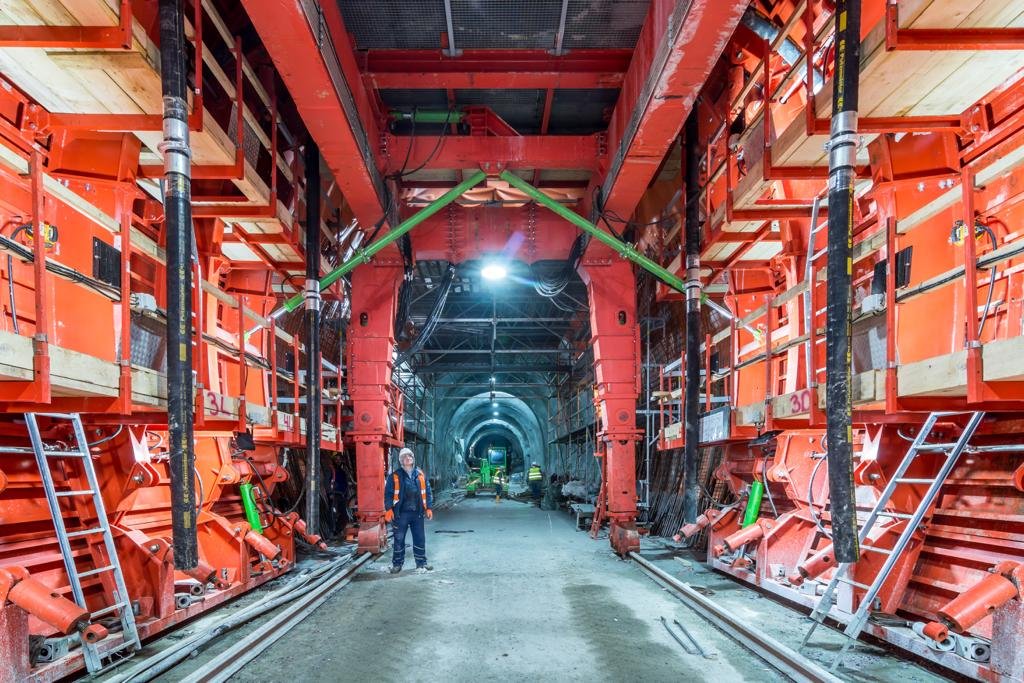 Underground tunnel construction with large red support structures, workers wearing helmets and safety gear, pipes, ladders, and equipment along the sides.