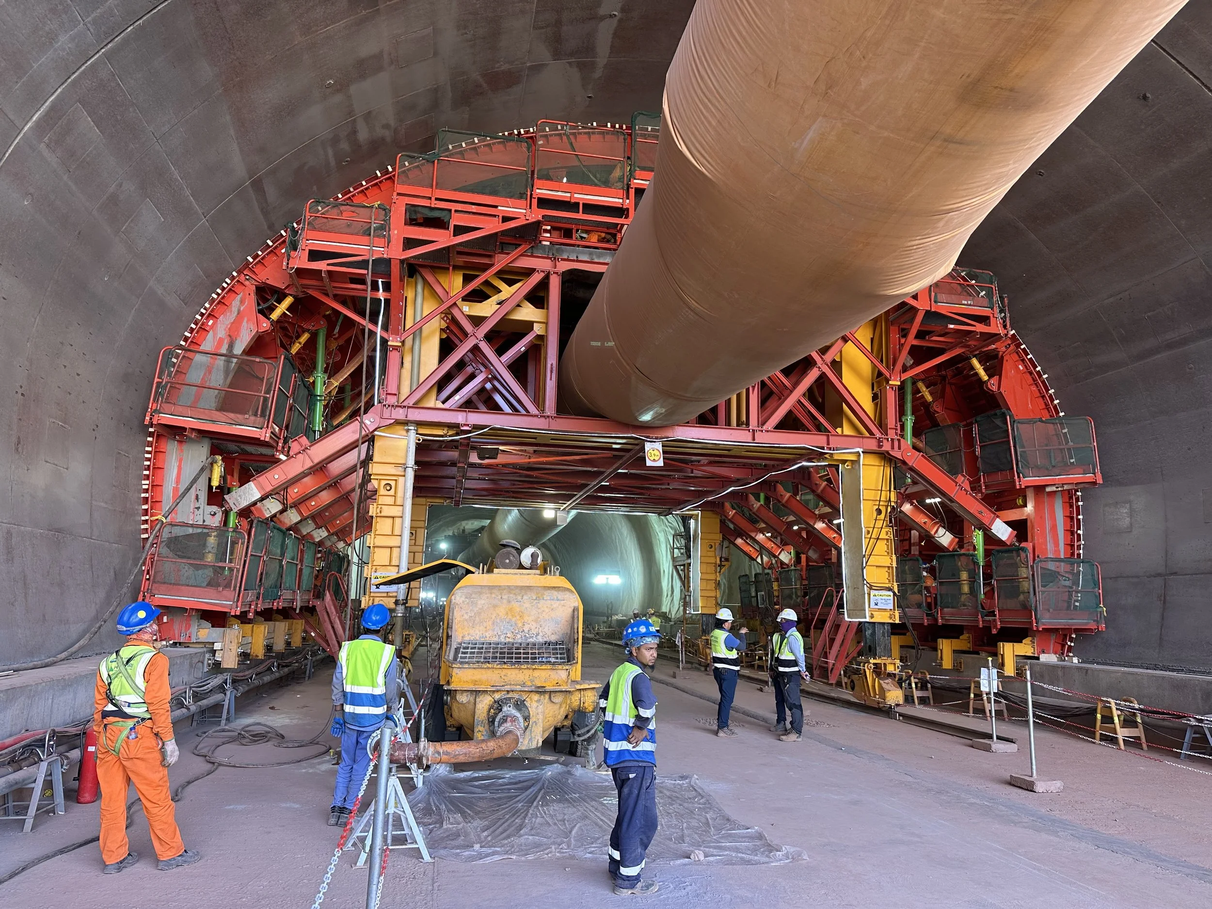 Construction workers in safety gear inspecting a large tunnel under construction with equipment and a giant pipe overhead.