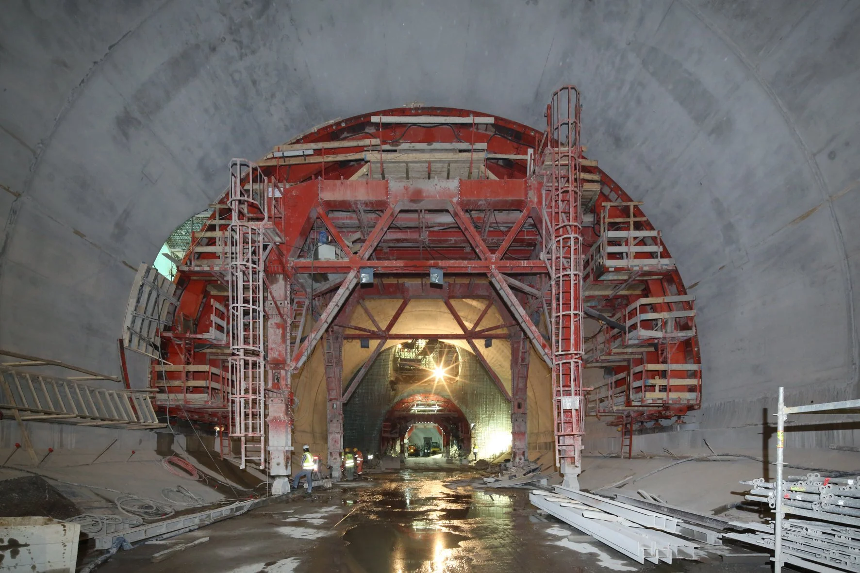 Underground tunnel under construction with workers and red scaffolding.