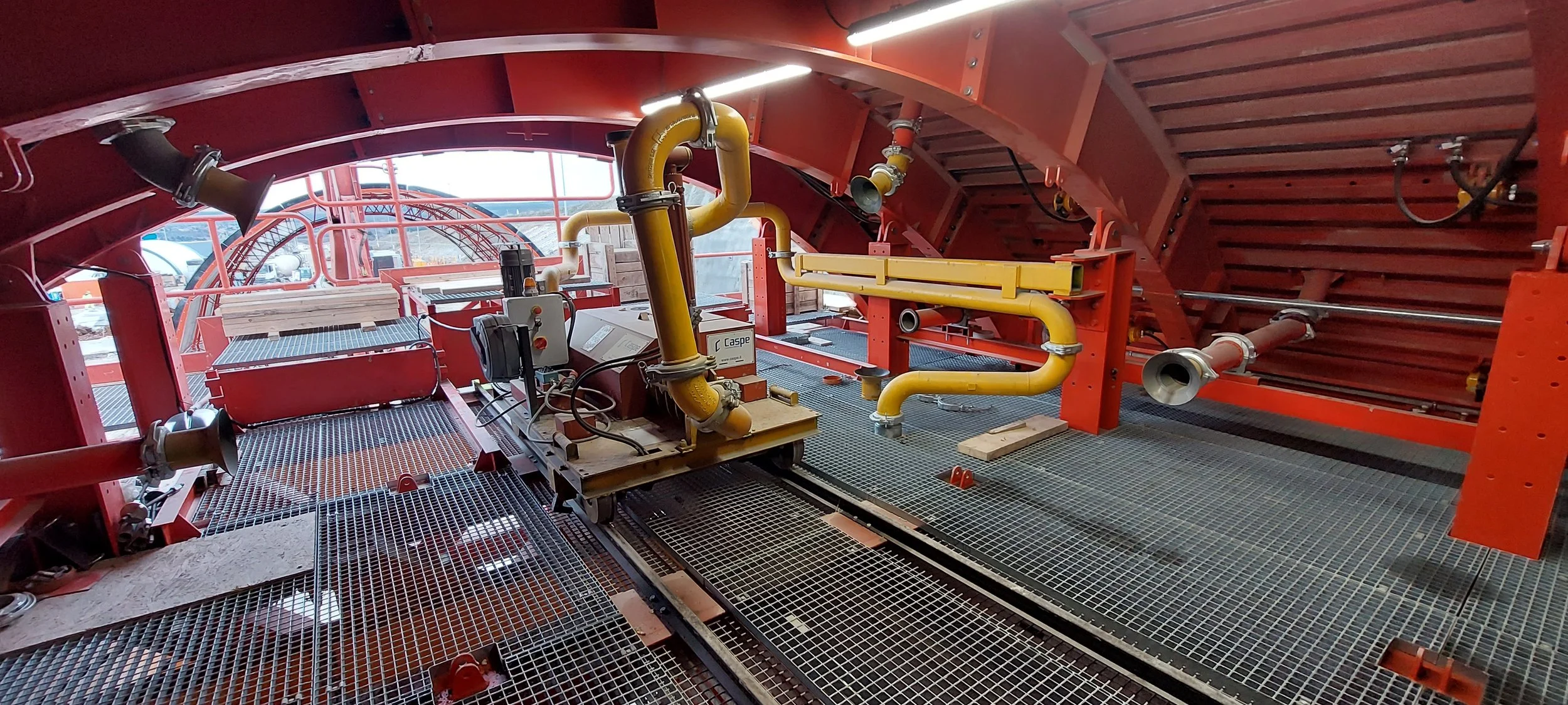 Construction site with orange metal structure, yellow pipes, and metal grating flooring, possibly part of an industrial or construction project.