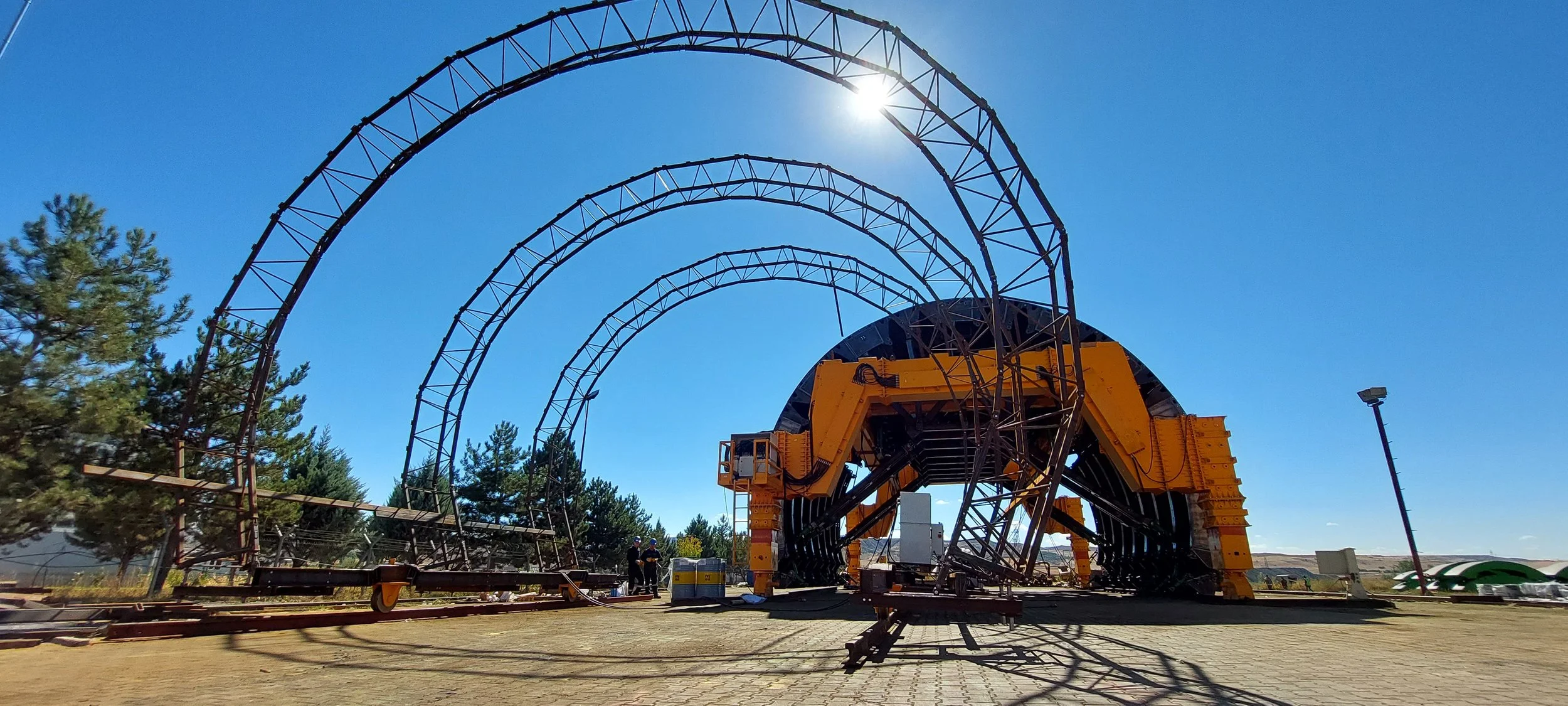 Large roller coaster track under construction with a yellow machine supporting a section of the ride, blue sky, trees, and construction workers visible.
