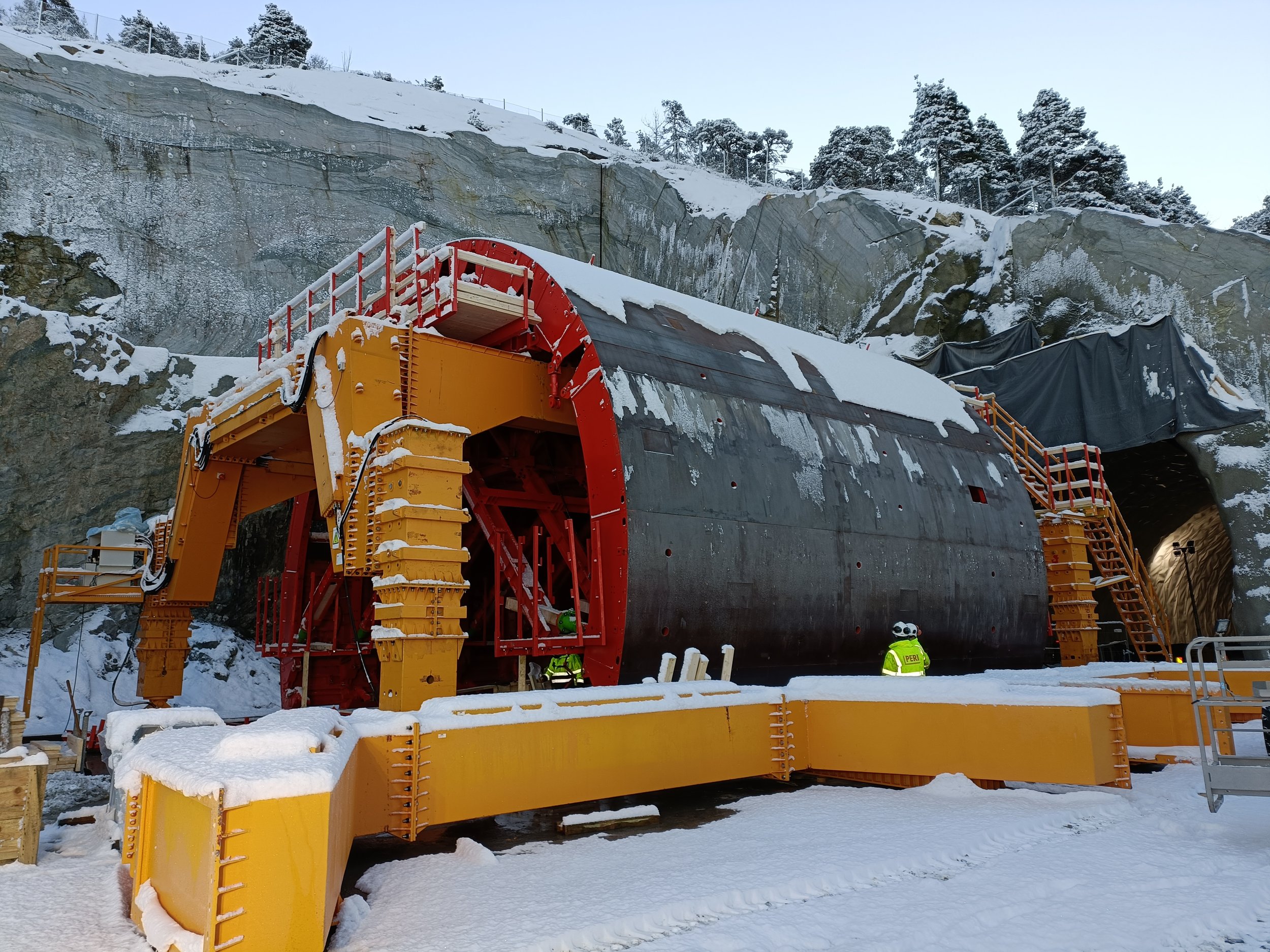 Construction workers in safety gear build a tunnel through a snowy mountain, with equipment and scaffolding around the entrance.