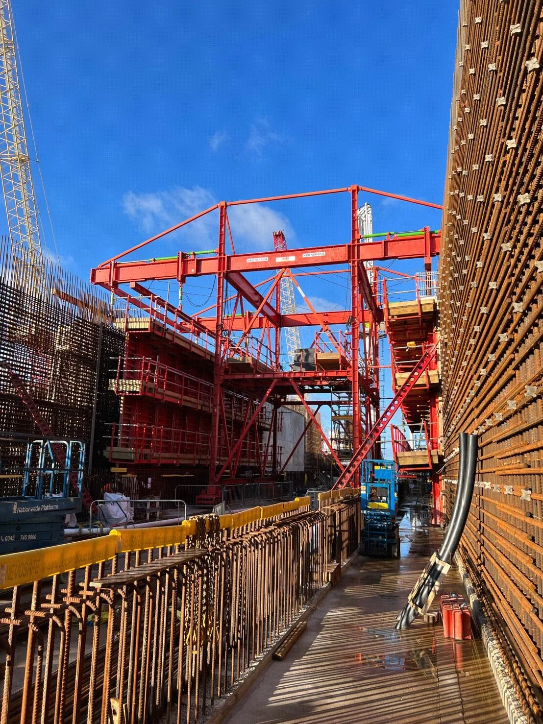 Construction site with red scaffolding, metal beams, and equipment, under a blue sky.