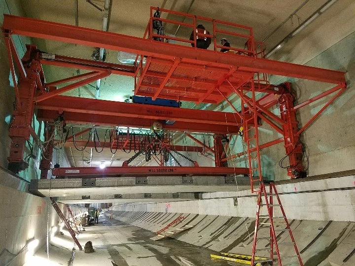 A large red construction gantry crane inside an underground tunnel, with workers on the upper platform and the crane lifting a large metal beam.
