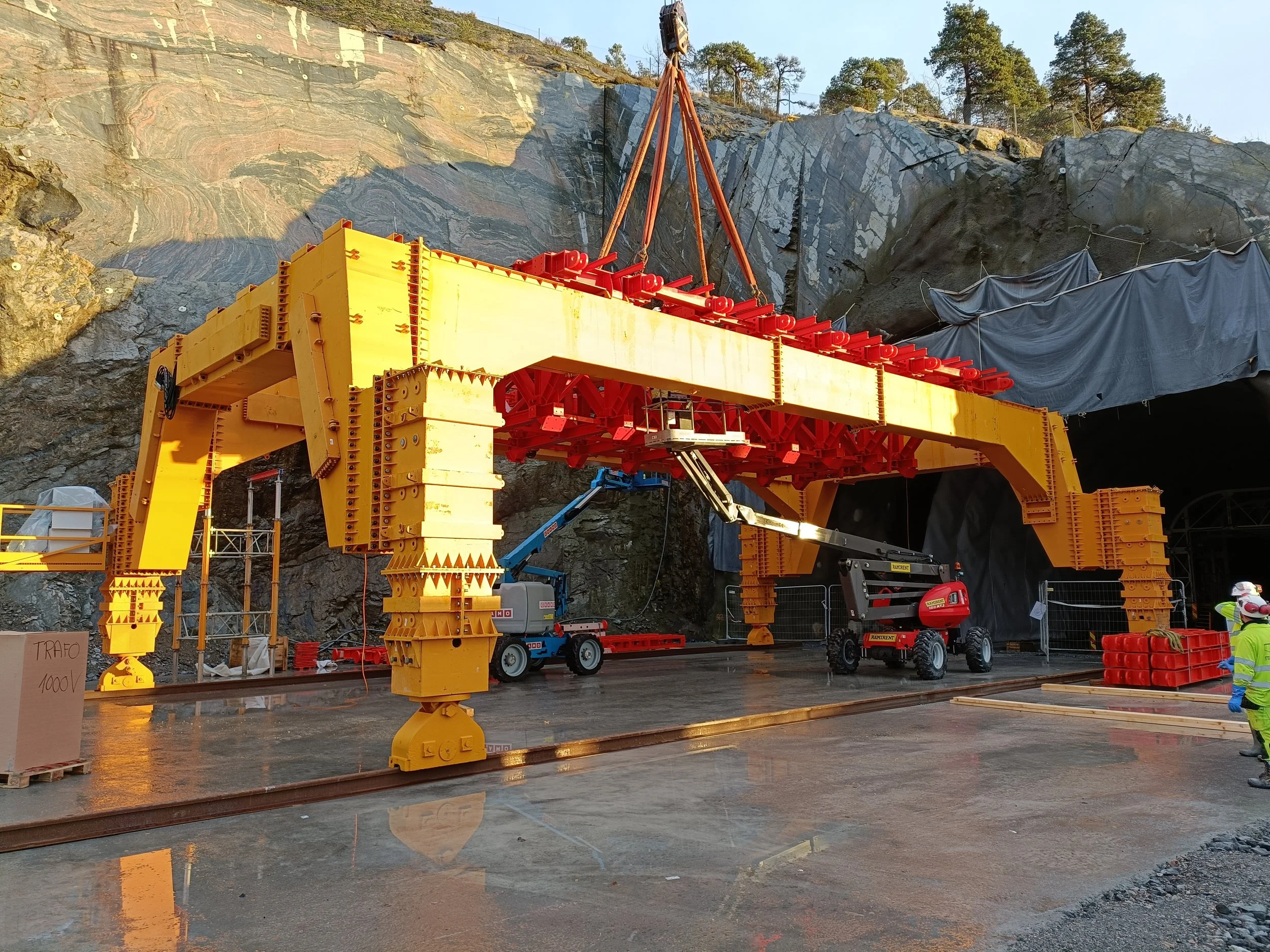 Construction site with large yellow and red tunnel boring machine being installed in a rocky hillside, with workers in protective gear observing.