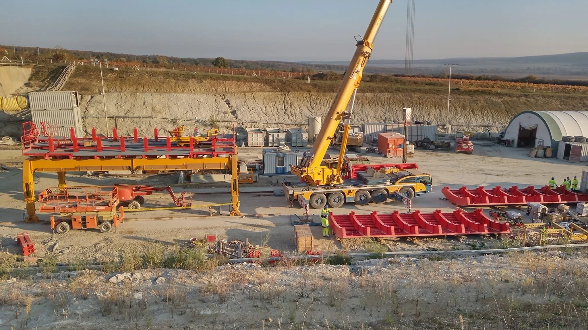 Construction site with workers, a crane lifting a large red structure, and several other construction vehicles and equipment in a rural area with fields and hills in the background.