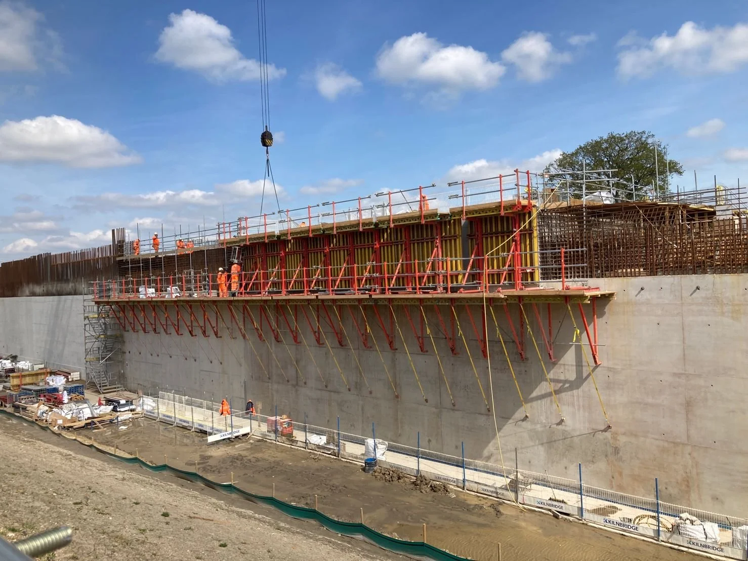 Construction workers in orange safety suits working on a large concrete structure with scaffolding and support beams, blue sky with clouds in the background.