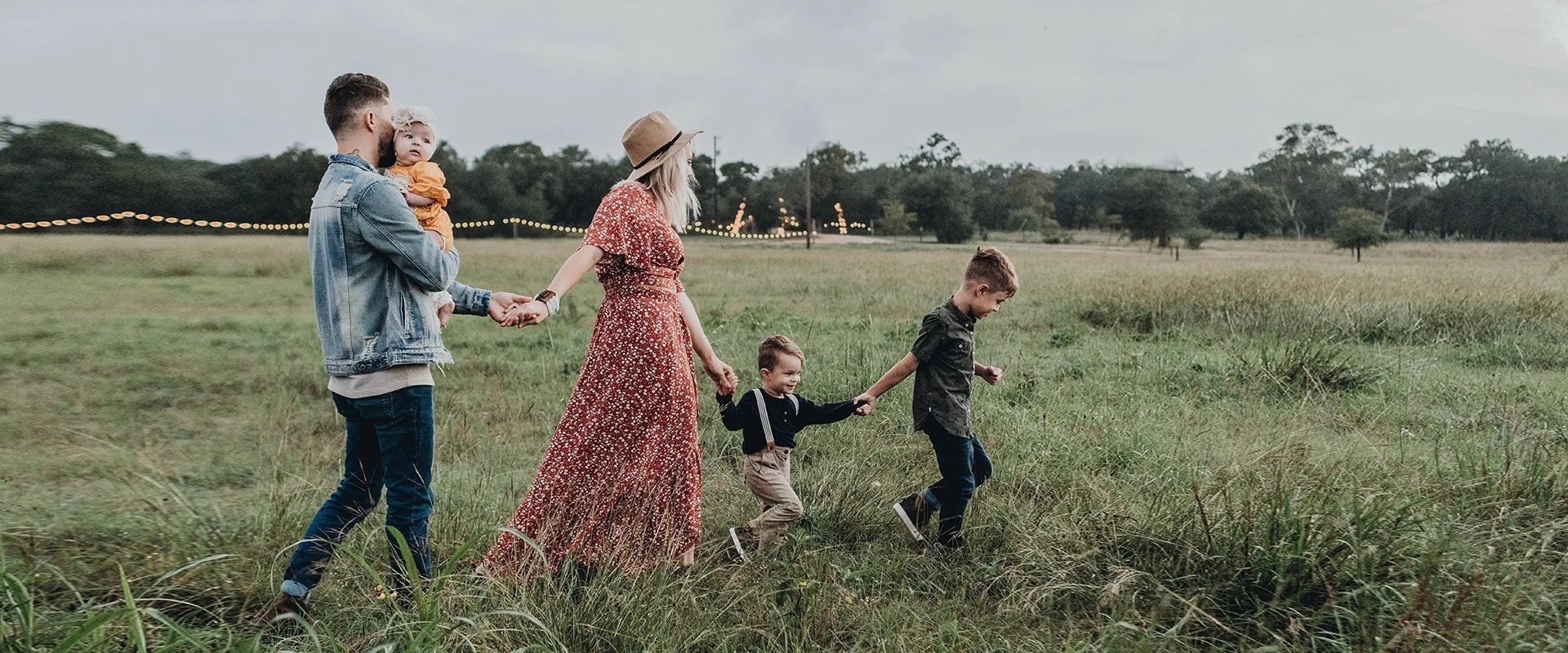 Familie beim Spaziergang in einem grünen Feld, Kinder halten an der Hand, Erwachsene halten sich an den Händen, im Hintergrund sind Bäume und Lichter zu sehen.