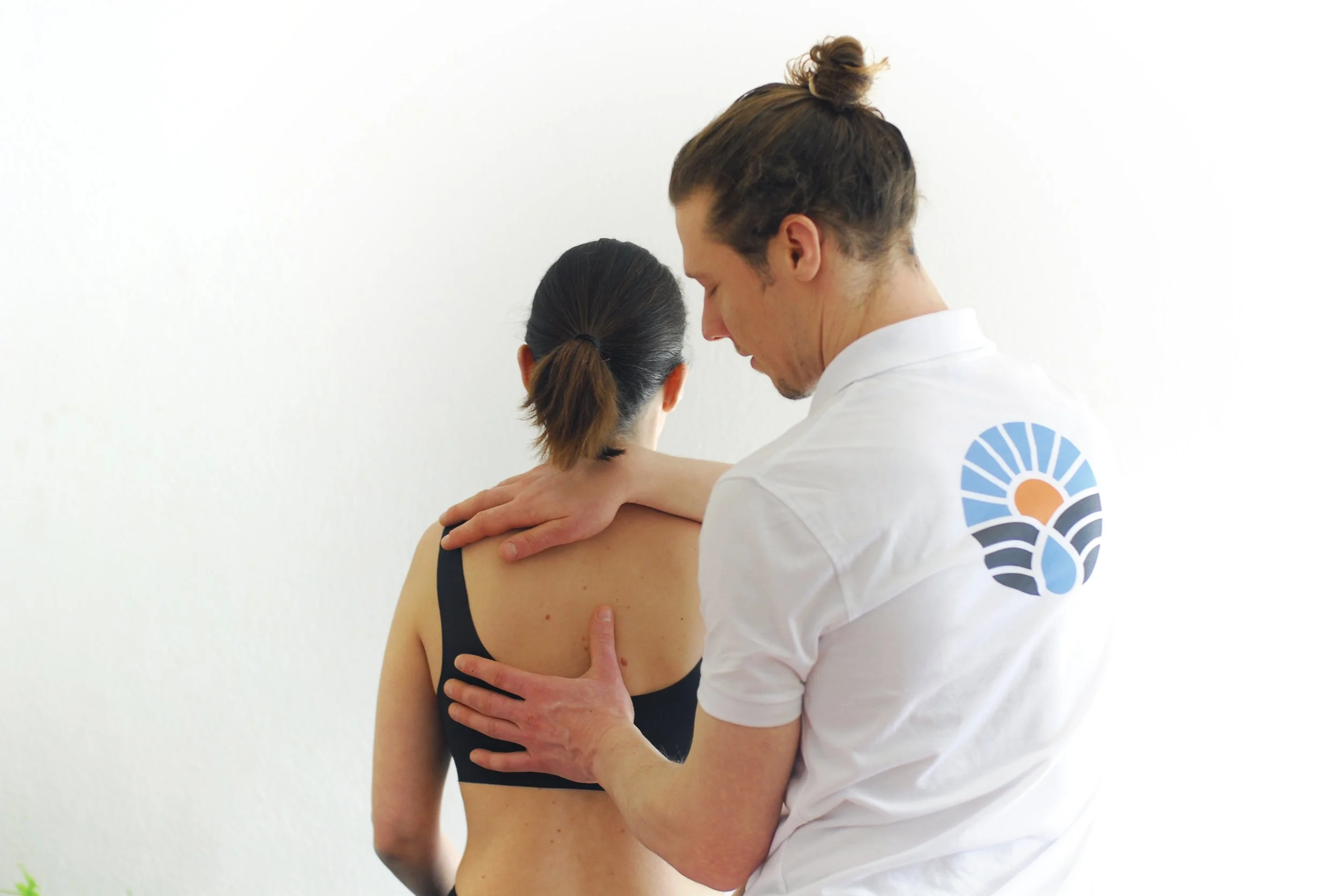 Ein Physiotherapeut führt eine Schulteruntersuchung bei einer jungen Frau durch, die schwarze Sportbekleidung trägt, vor einem weißen Hintergrund.