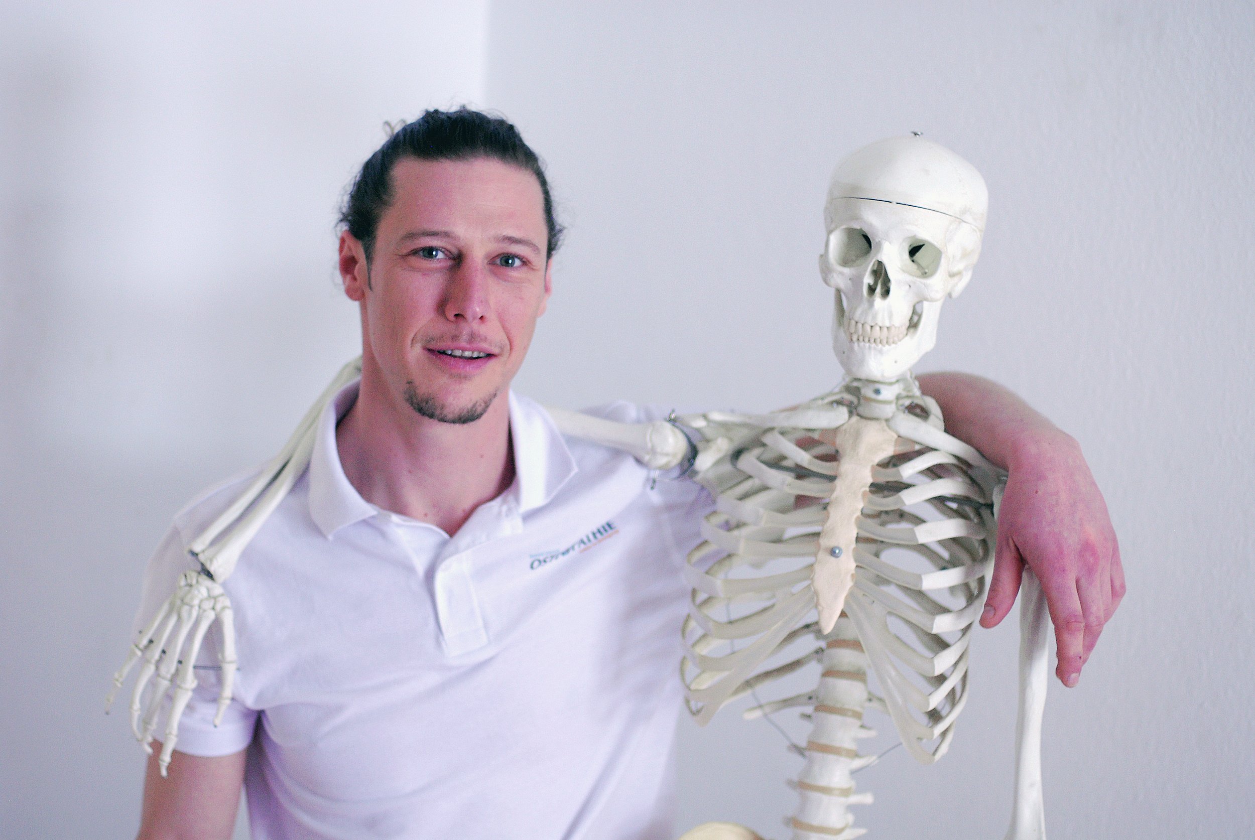 Osteopath Daniel Görich mit dunklem Haar und weißem Hemd steht neben einem menschlichen Skelett und legt den Arm über die Schulter des Skeletts.