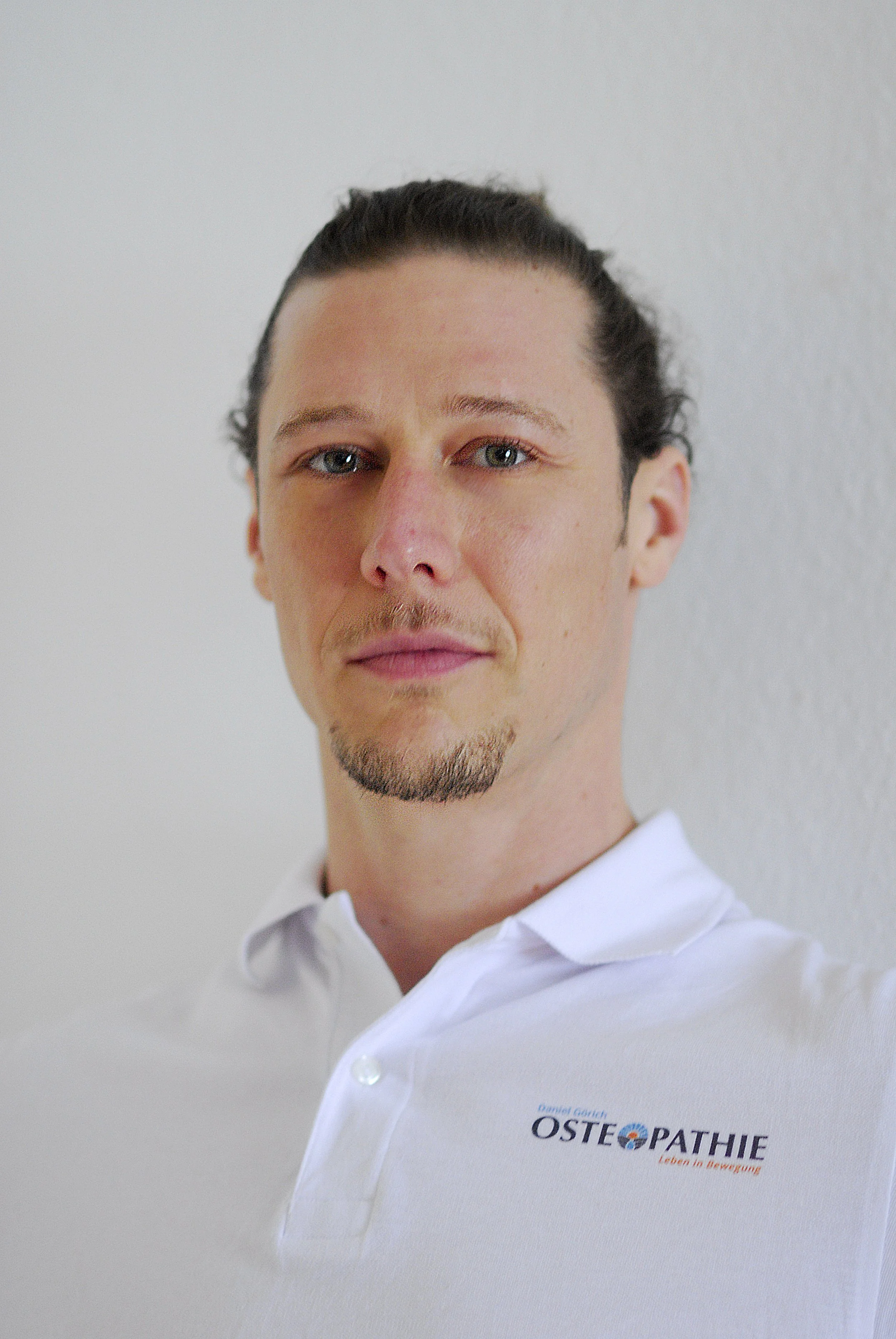Porträt von Daniel Görich mit dunklen, gesunden Haaren, gebürstetem Bart, weißem Hemd mit Logo auf der linken Brust, steht vor weißer Wand.