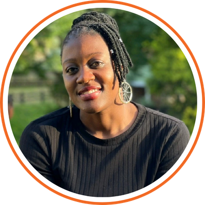 Portrait of a smiling woman with short dreadlocks, wearing earrings and a black top, outdoors with green foliage in the background.