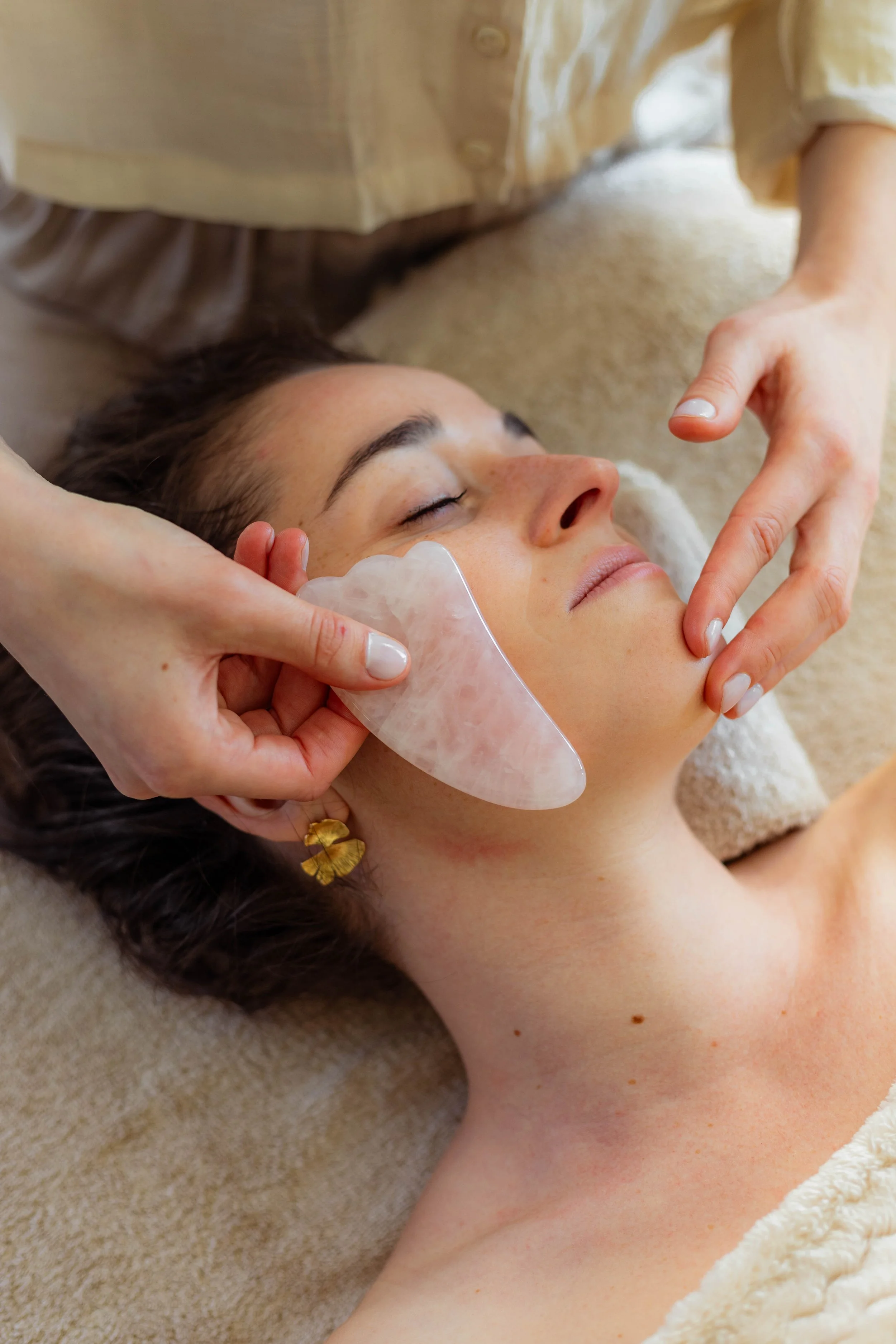 Une femme reçoit un traitement facial avec un galet en pierre rose, allongée sur une table de massage. La praticienne utilise ses mains pour appliquer le soin.