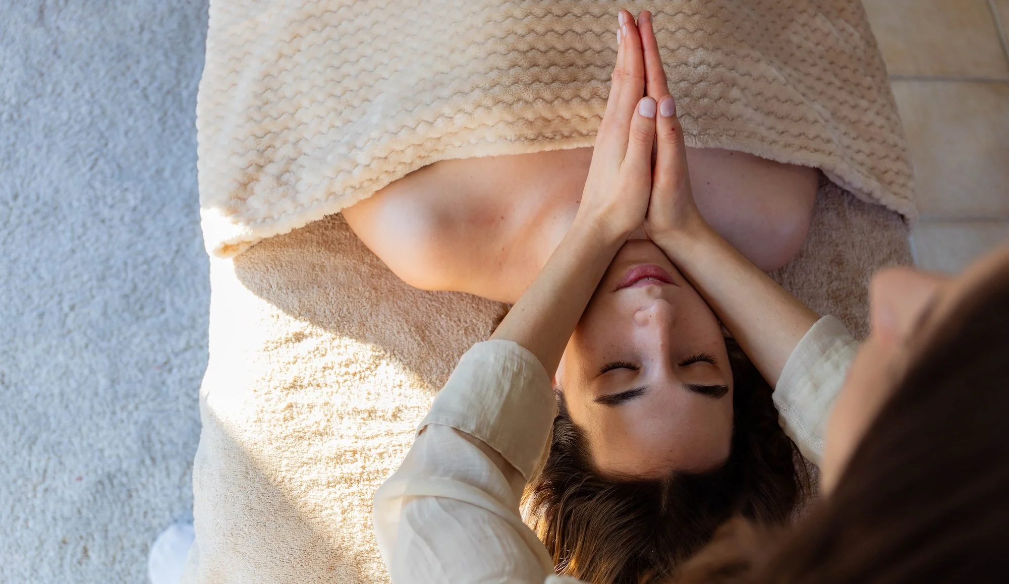 Une femme reçoit un massage avec les mains de l'autre personne appuyées sur ses épaules, sur un lit de massage recouvert d'une serviette beige