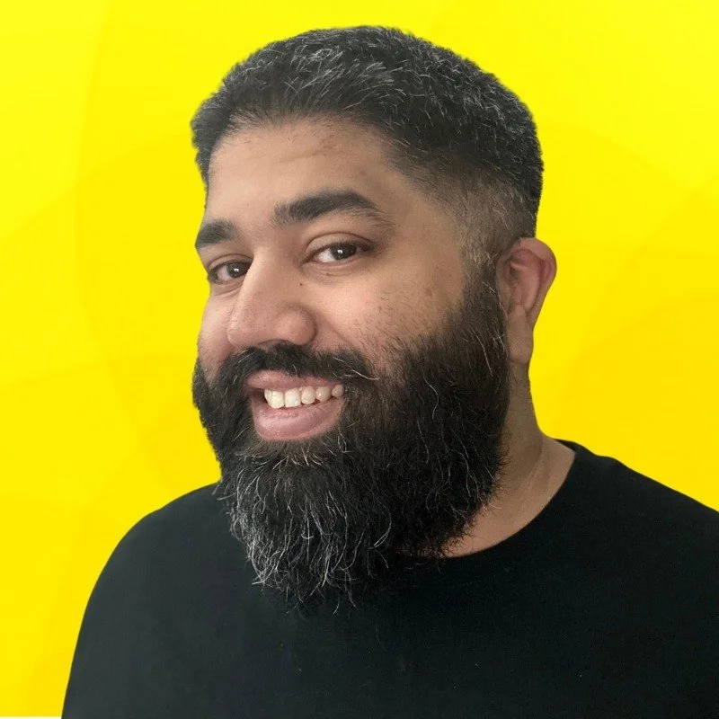 A man with a dark beard and short dark hair, smiling, wearing a black shirt against a bright yellow background.