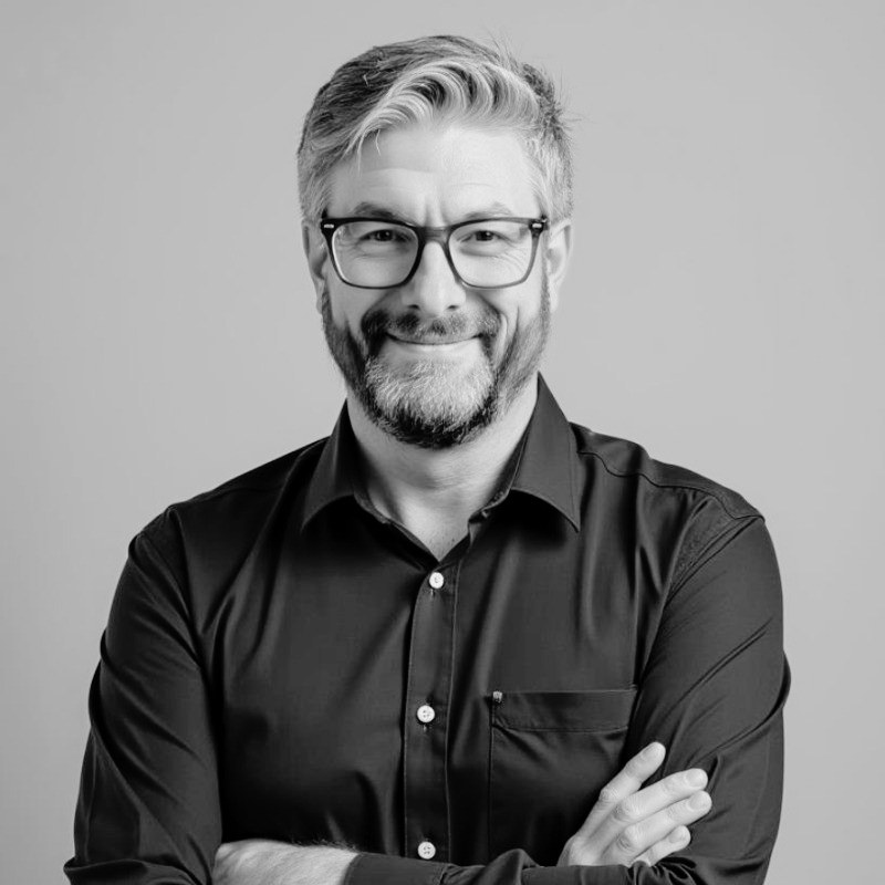 Portrait of a man with glasses, a beard, and a smile, wearing a dark button-up shirt against a plain background.