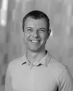 A professional man with short hair smiling, wearing a collared shirt, standing in front of a blurred background.