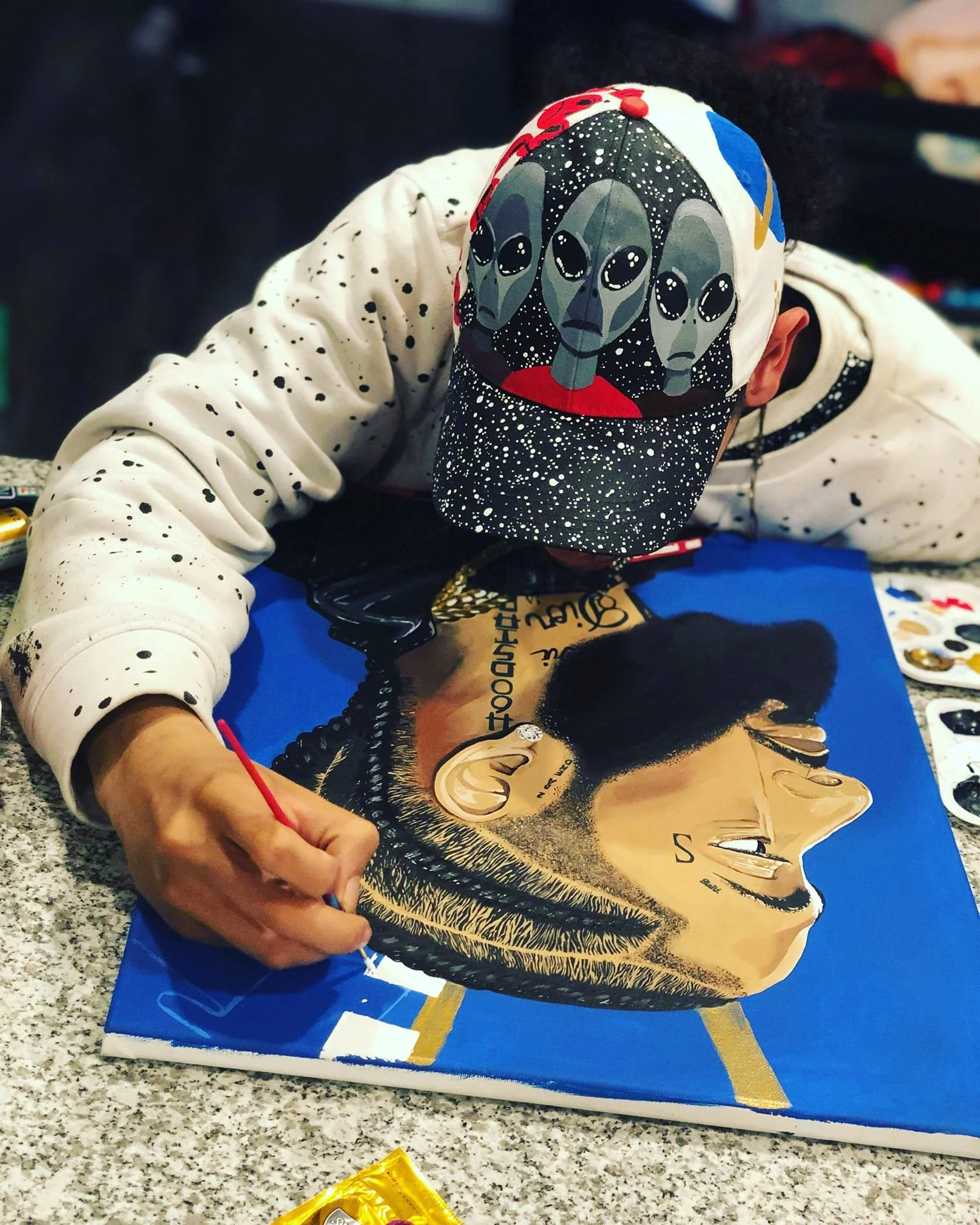 Artist painting a portrait of a man with a beard on a blue canvas. The artist is wearing a cap with painted faces and a white shirt with black spots, working on detailed features of the portrait.