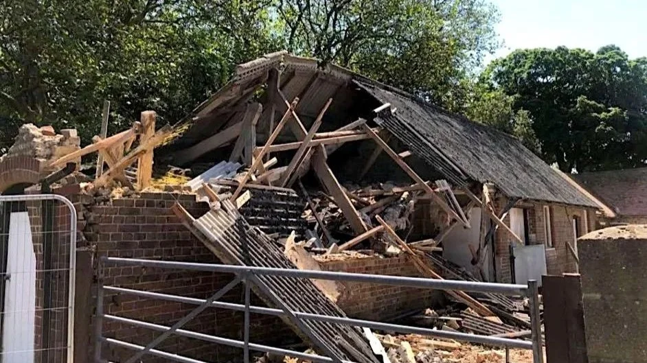 A partially collapsed brick house with the roof caved in and debris scattered around, including broken wood and other materials, behind a metal fence. outbuilding or barn ready for demolition. containing asbestos