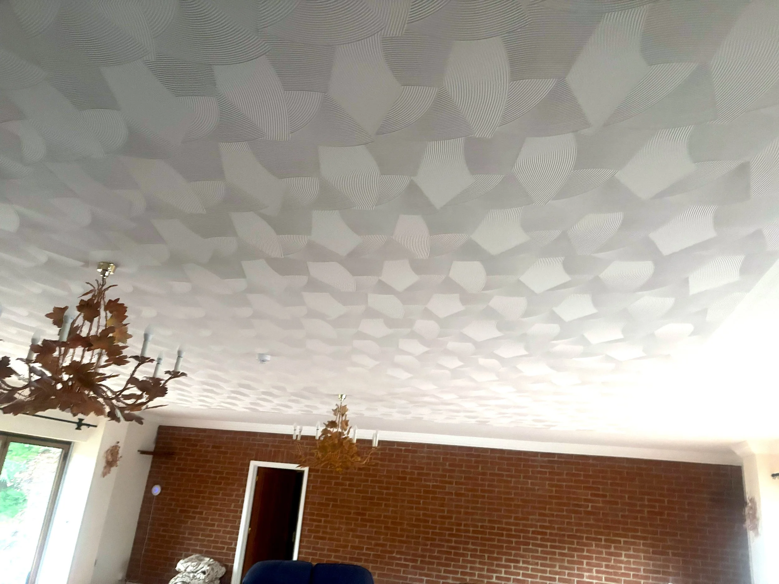 Living room with a white textured ceiling containing asbestos, two leaf-shaped chandeliers, a brick accent wall, and a window on the left side. 