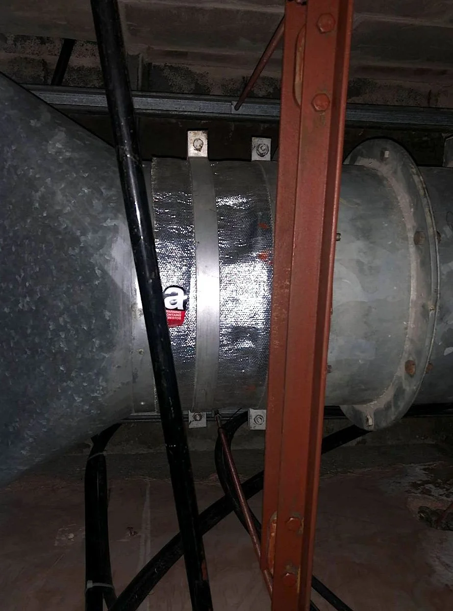 A large, cylindrical, metal HVAC duct connected to a round metal vent containing asbestos. Signs of oxidation and corrosion, with visible rust and wear. It is secured with metal brackets and is situated in a ceiling or attic space with wooden beams.