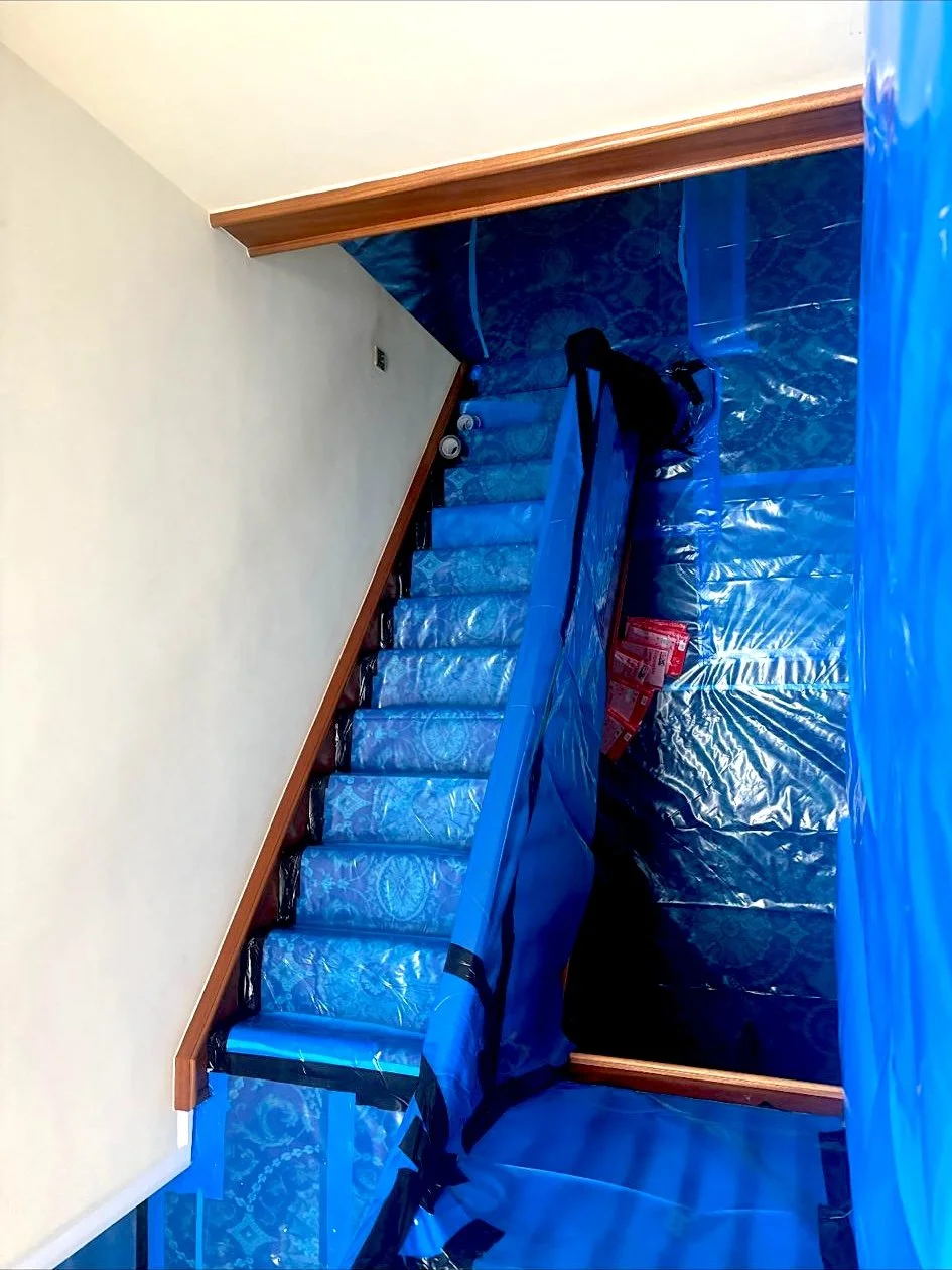 Hallway ceiling containing asbestos. Asbestos removal in process. Blue plastic covering to protect the staircase and floor, with a black backpack and cans on stairs, indicating construction or renovation work.