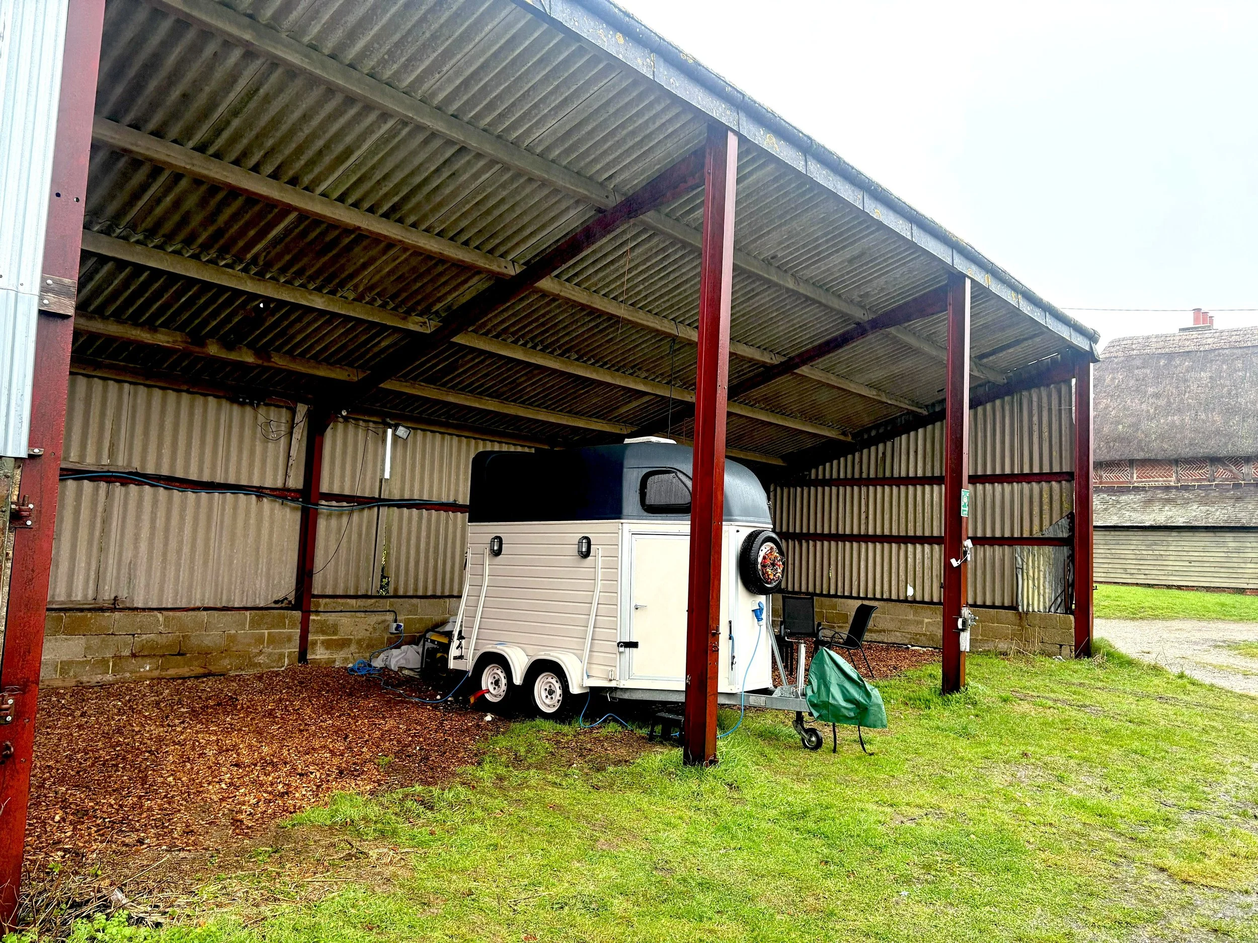 Small white trailer with black top parked under a metal barn or outbuilding with wooden beams, green grass around, and a brick wall in the background. The building contains asbestos needing removal and management.