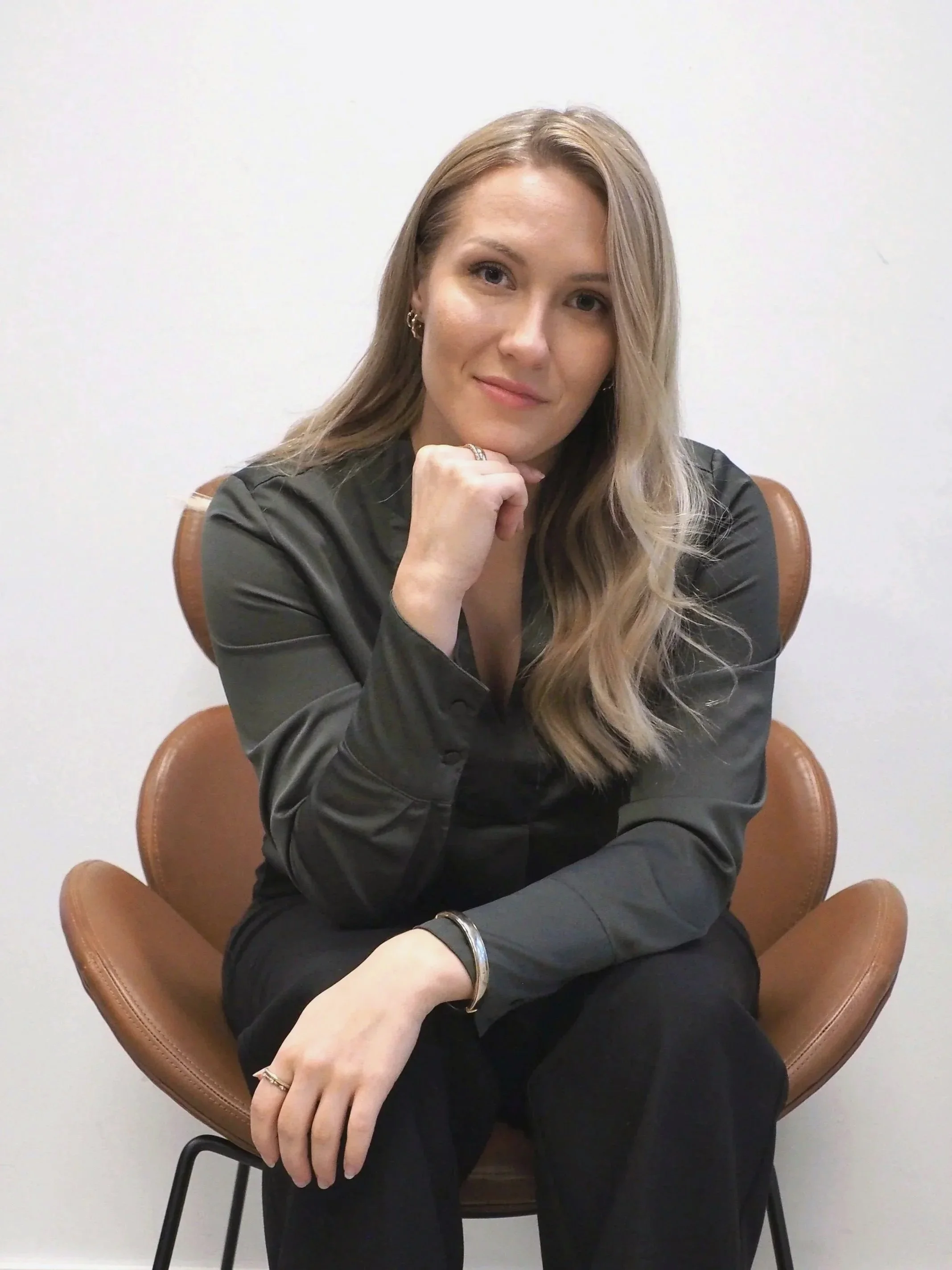 A woman with long blonde hair, wearing a dark green blouse and black pants, sitting on a brown chair with a white wall background, resting her chin on her hand and smiling.