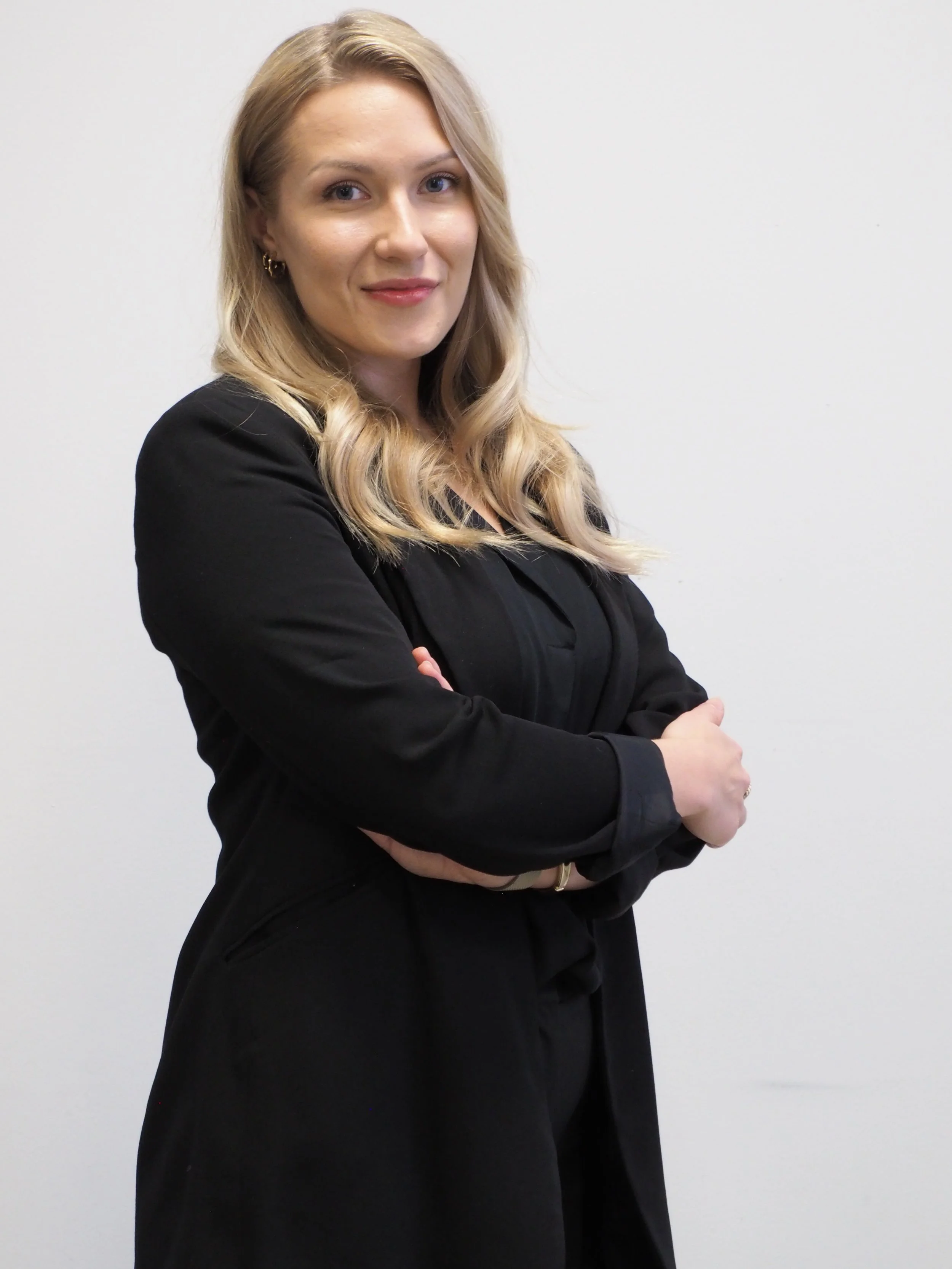 A woman with long blonde hair wearing a black blazer and black pants, standing against a plain white wall, with arms crossed and a slight smile.
