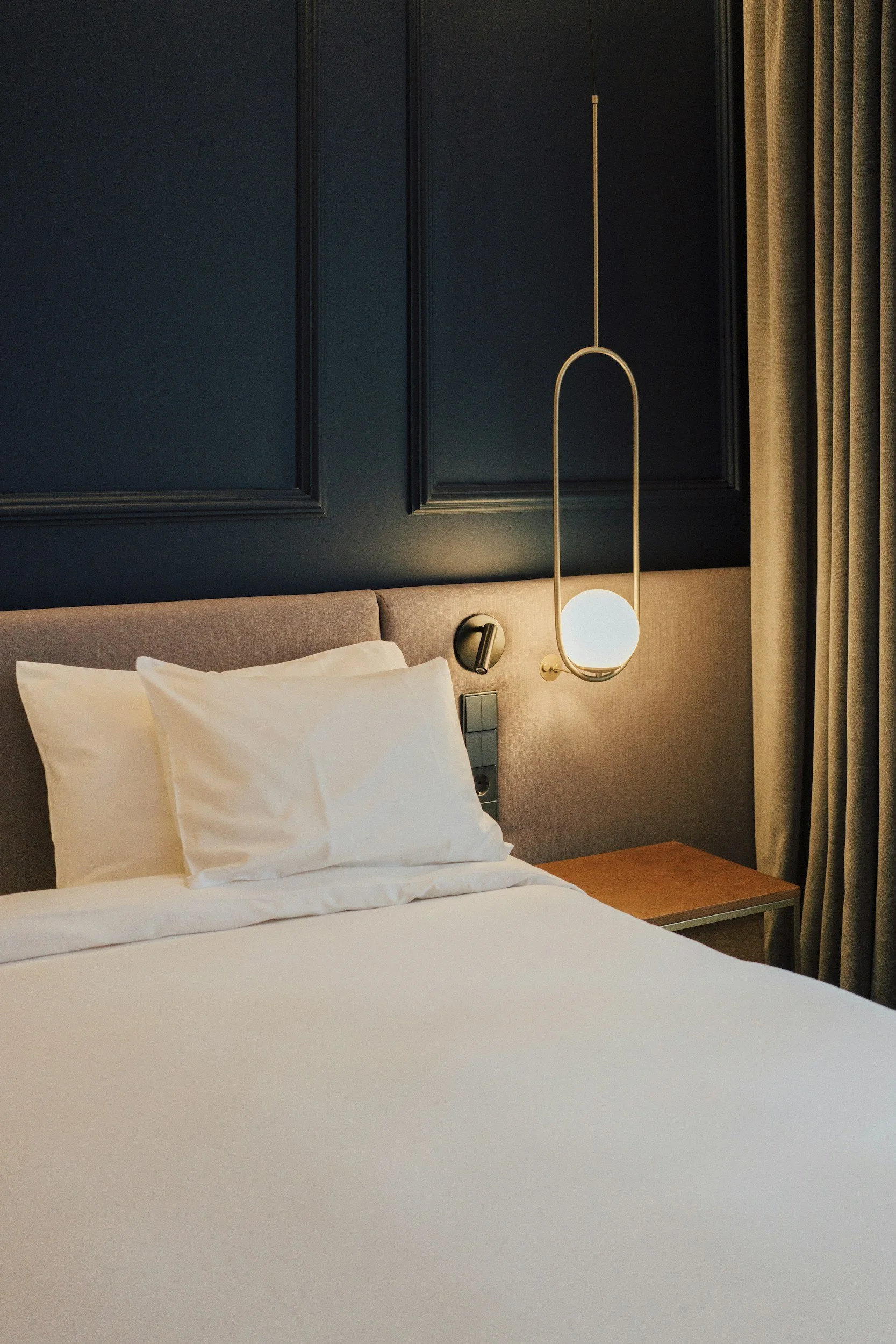 Un lit avec deux oreillers blancs dans une chambre avec un mur bleu foncé, une lampe suspendue moderne et des rideaux beige.