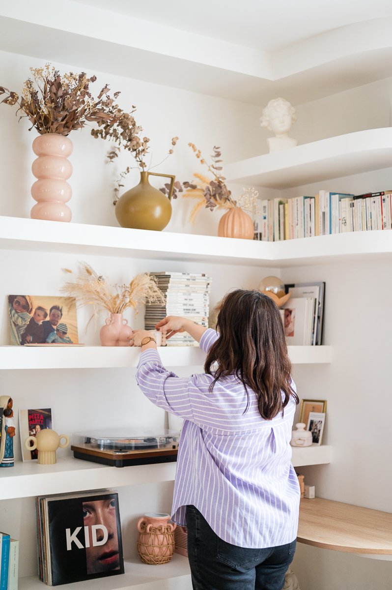 Une jeune fille avec des cheveux bruns longs, portant une chemise à rayures violettes et blanches, qui arrange des objets sur une étagère blanche dans un intérieur moderne avec des décorations en poteries, des livres et des photos.