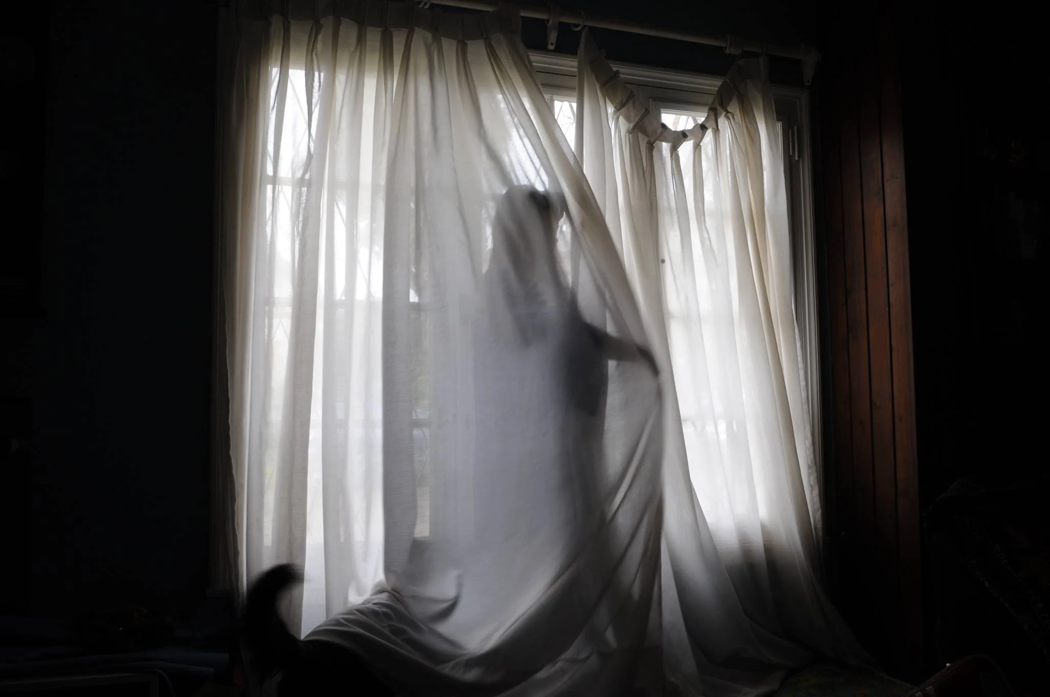 A silhouette of a person behind a sheer curtain, standing near a window with white curtains, in a darkened room.
