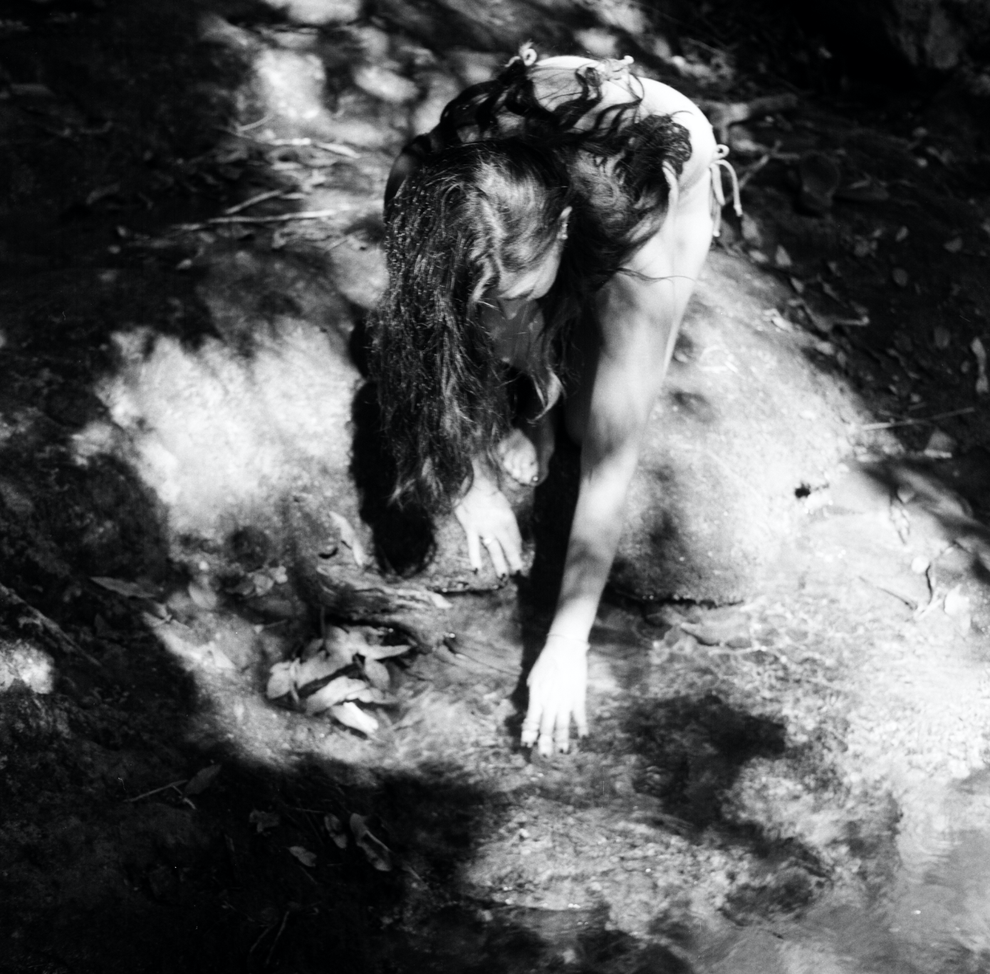 Black and white photo of a woman kneeling near a stream in a natural setting, with her hair wet and her hand in the water.