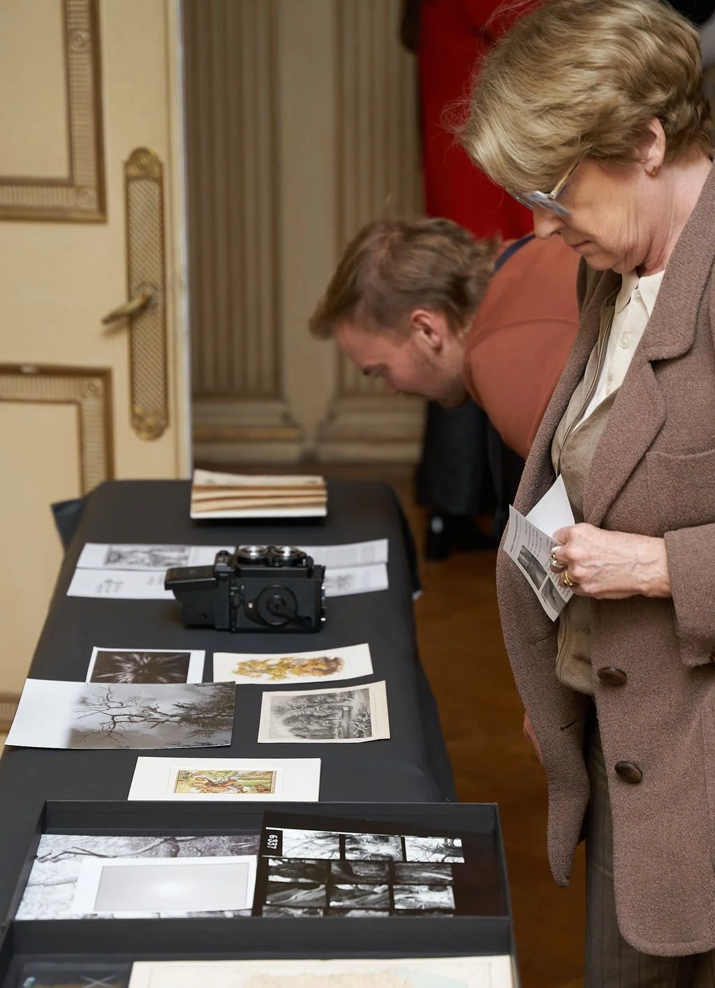 Two women and a man looking at photographs and artwork arranged on a table, with a camera among the displayed items, in an elegant room with ornate decor.