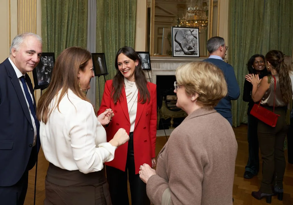 A group of people socializing in an elegant room with green curtains and framed artwork, including a fireplace and a chandelier.