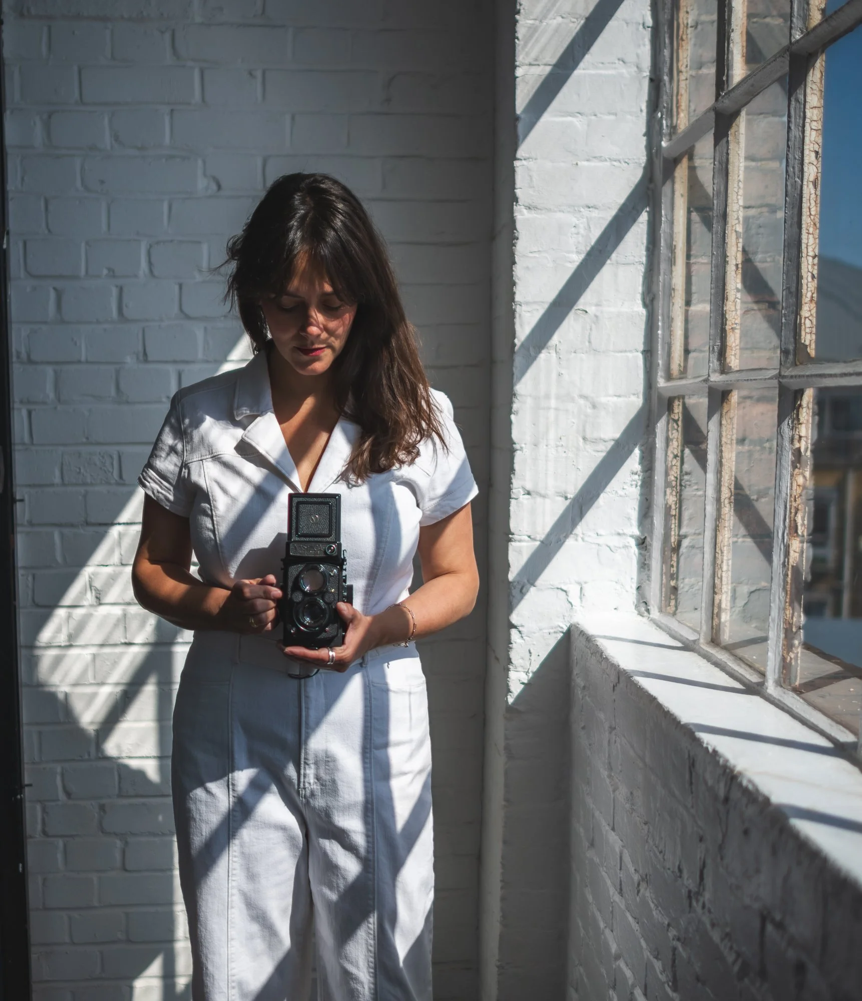 A woman in an all-white outfit holding a vintage camera in front of a white brick wall and window with shadows cast by sunlight.