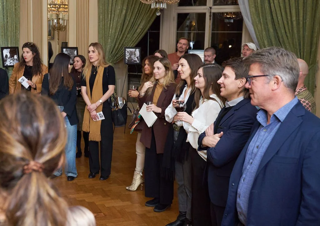 Group of people at a social event listening to a speaker, some holding wine glasses, in an elegant room with framed photographs and green curtains.