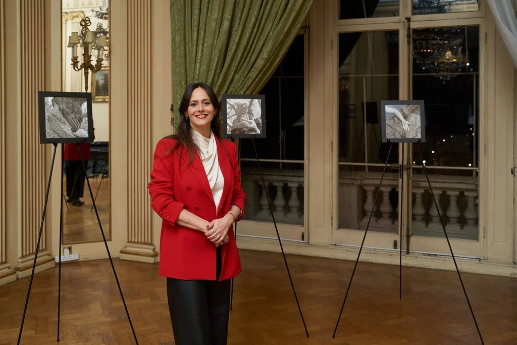 A woman in a red blazer and black pants standing in an art gallery, smiling at the camera, with black and white photographs on easels behind her in a well-decorated room.