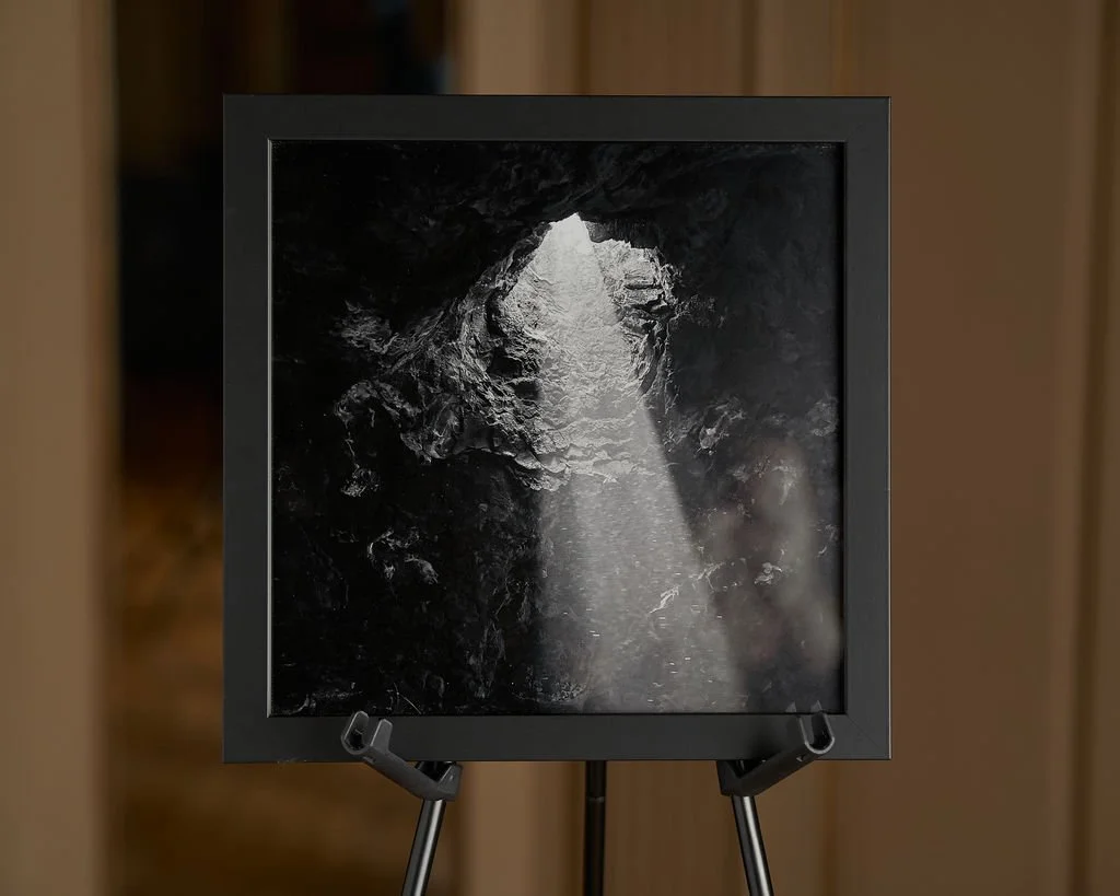 A framed black and white photograph or artwork depicting a rocky cave with light streaming in from an opening above.