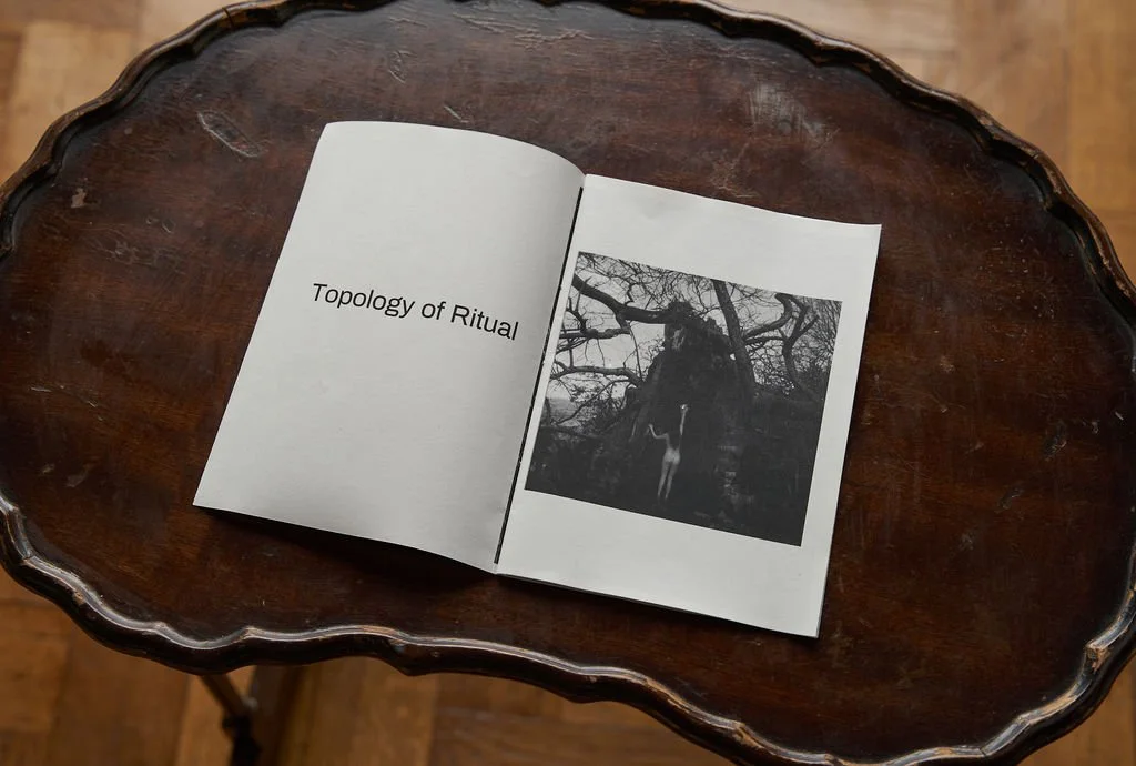 Open booklet on wooden table titled "Topology of Ritual," with black and white photo of a person hanging from a tree.