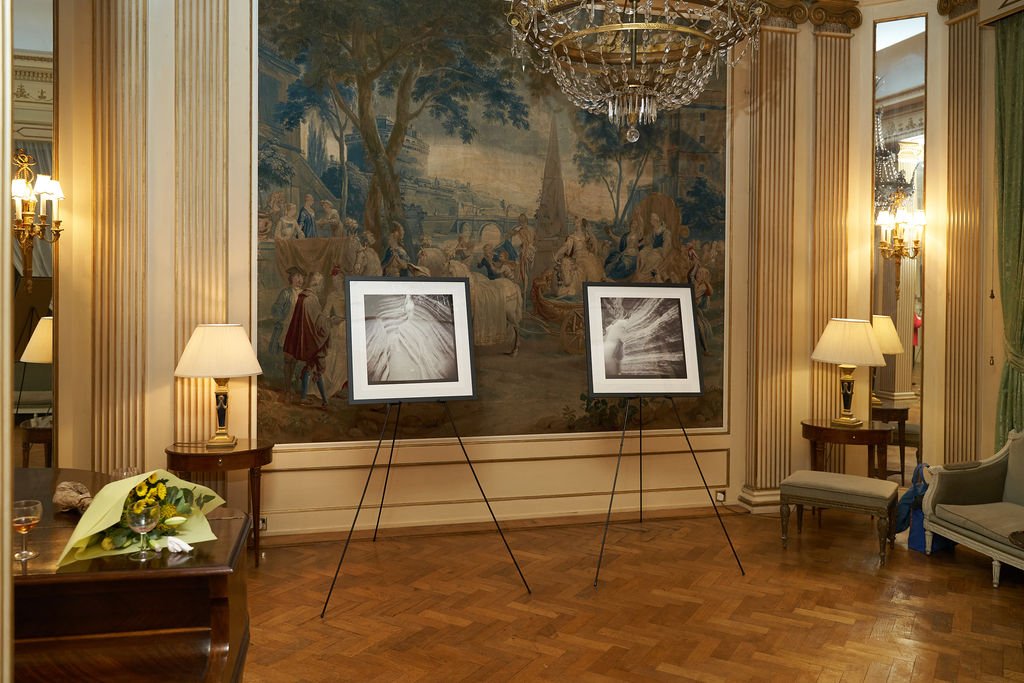 An elegant room with a large classical mural on the wall, two framed black-and-white photographs on easels, side tables with lamps, and furniture including chairs and a table with a bouquet of flowers.