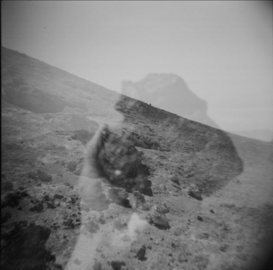 Black and white image showing a rocky, barren landscape with a large mountain or hill in the background, and a reflection of a person holding a camera in the foreground.