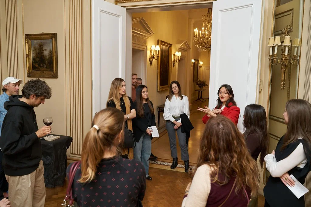 A woman in a red blazer giving a talk or tour to a small group in an elegant, well-lit room with ornate decorations and chandeliers.