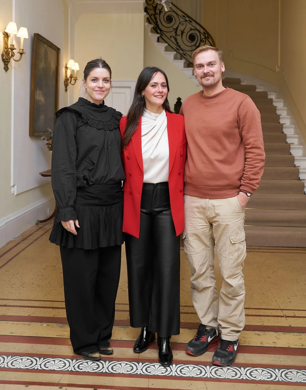 Three people standing together in a stairwell, smiling at the camera. The woman on the left is wearing a black dress with ruffles, the woman in the middle is wearing a red blazer over a white blouse and black pants, and the man on the right is wearin