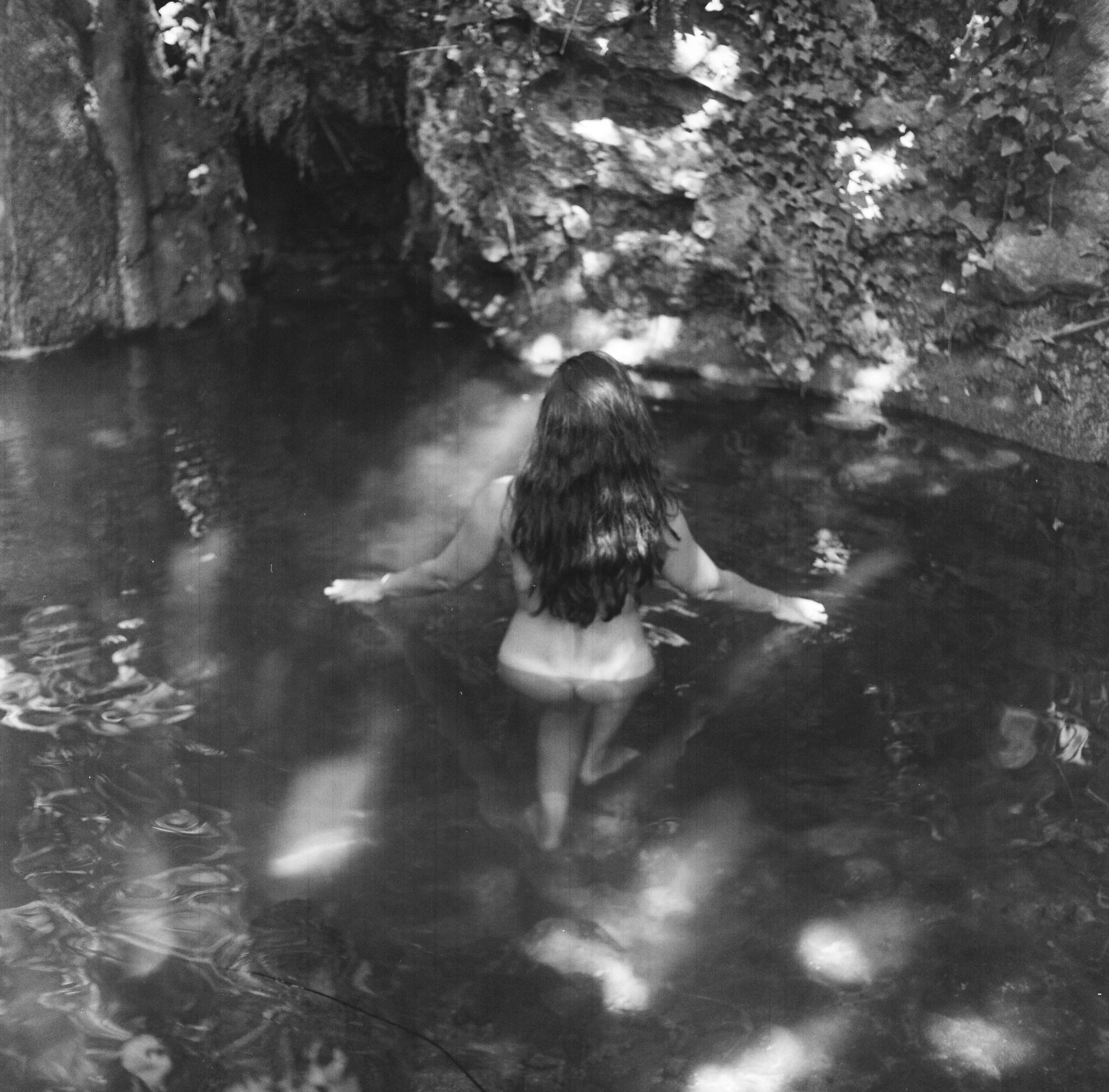 A woman with long hair, seen from the back, standing in a natural water pool surrounded by rocks and foliage.